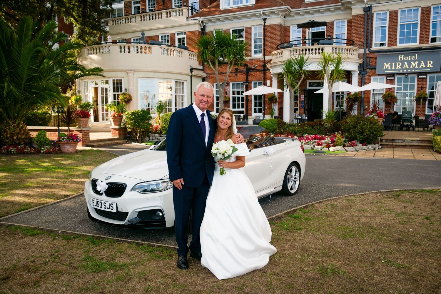 Bride and groom pose beside a white BMW convertible outside Hotel Miramar on their wedding day.