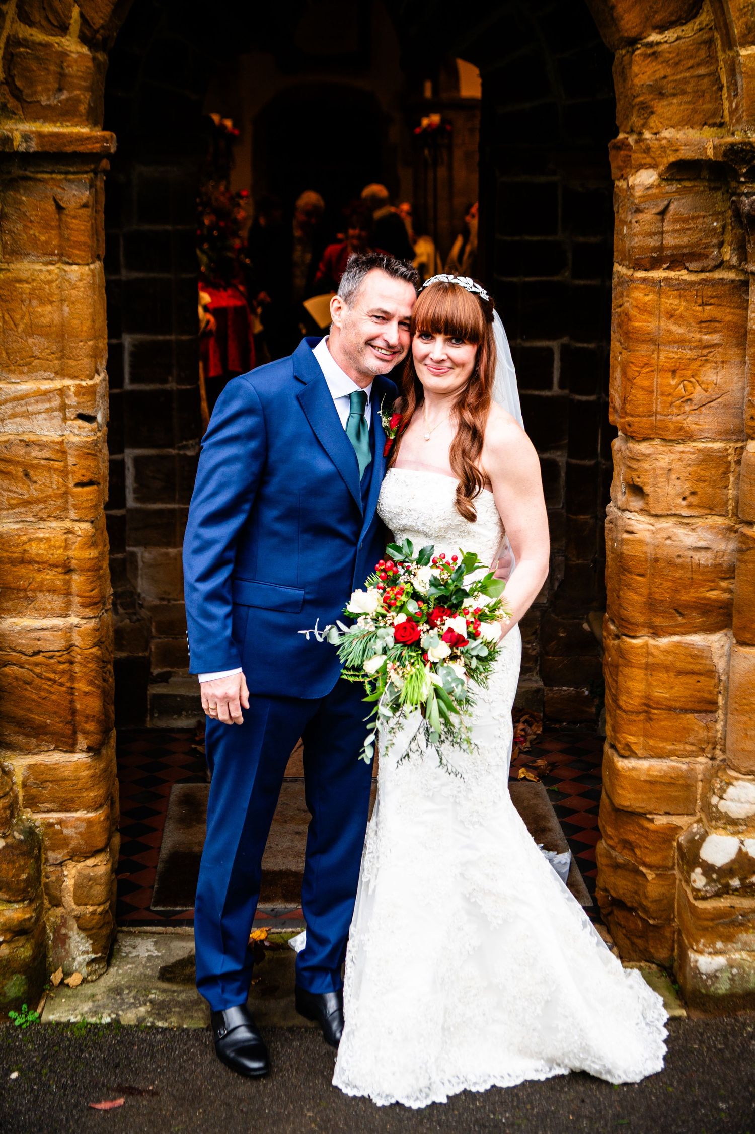 Bride and groom posing together at a stone church doorway, bride holding red and white bouquet.