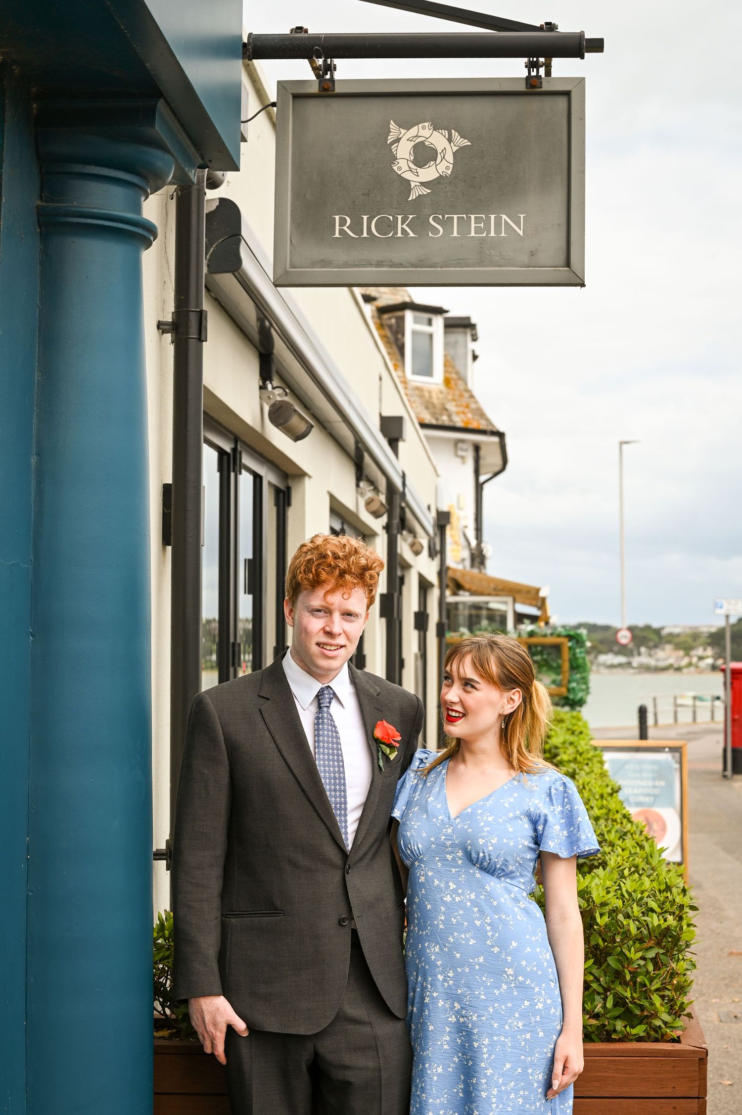 Couple posing outside Rick Stein restaurant, man in suit with red boutonniere, woman in blue floral dress, waterfront setting.