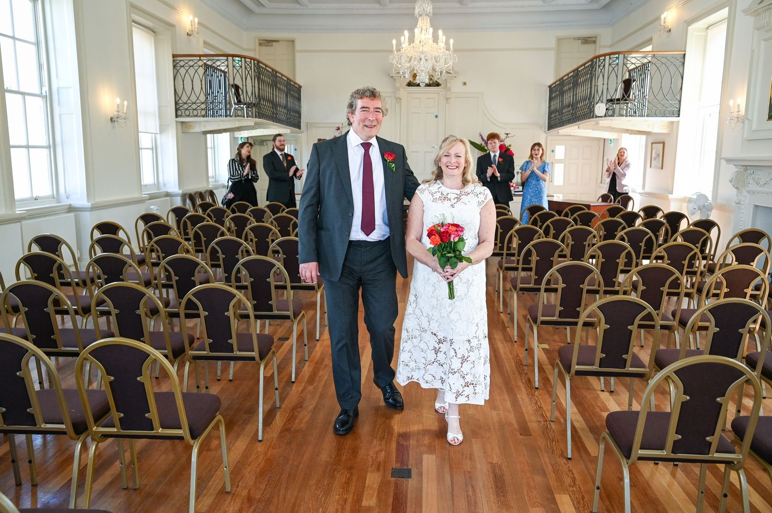 Couple walks down the aisle of an elegant wedding venue with rows of chairs and a chandelier.