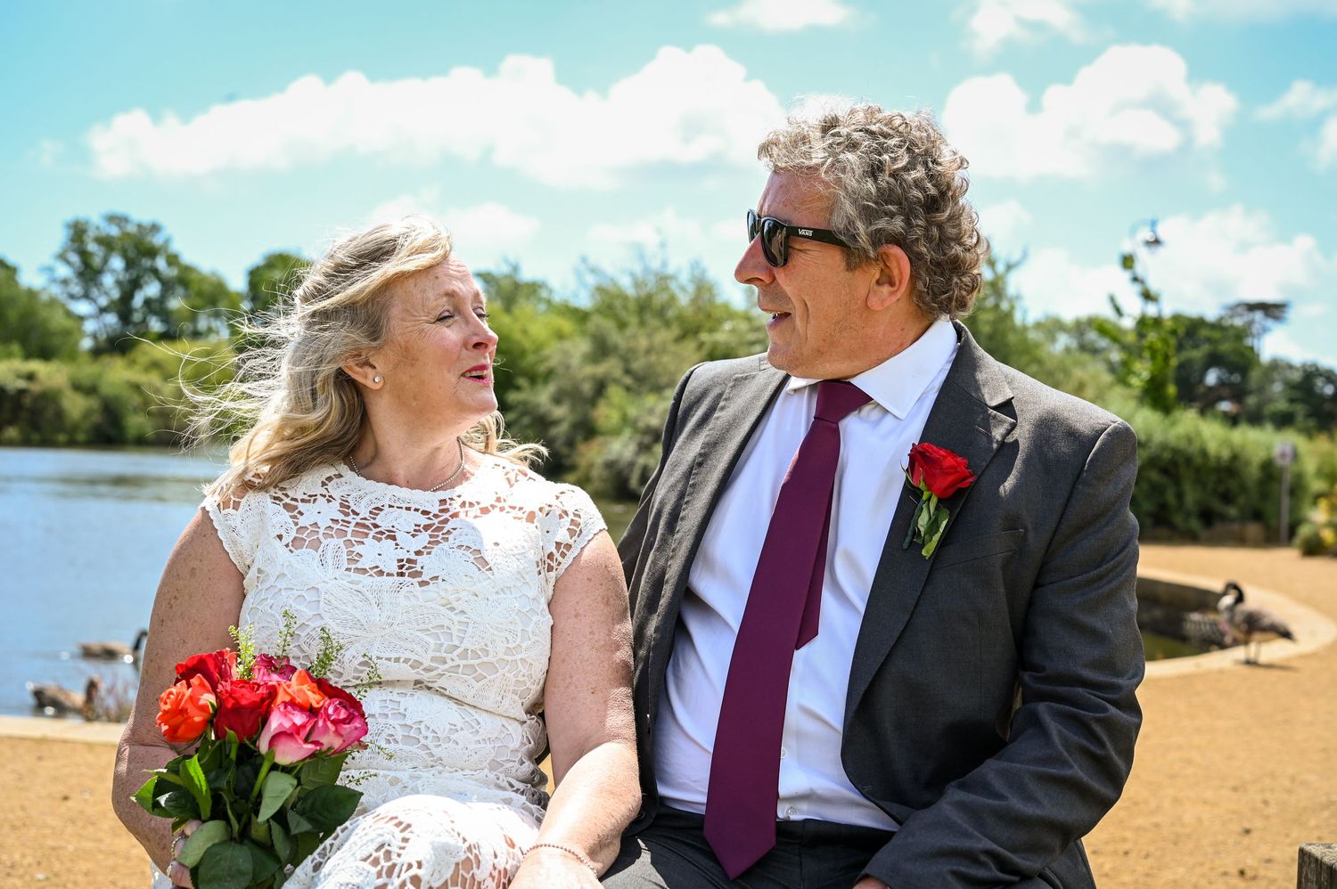 Couple in wedding attire smiling at each other outdoors, bride holding pink roses, groom wearing red tie and boutonniere.
