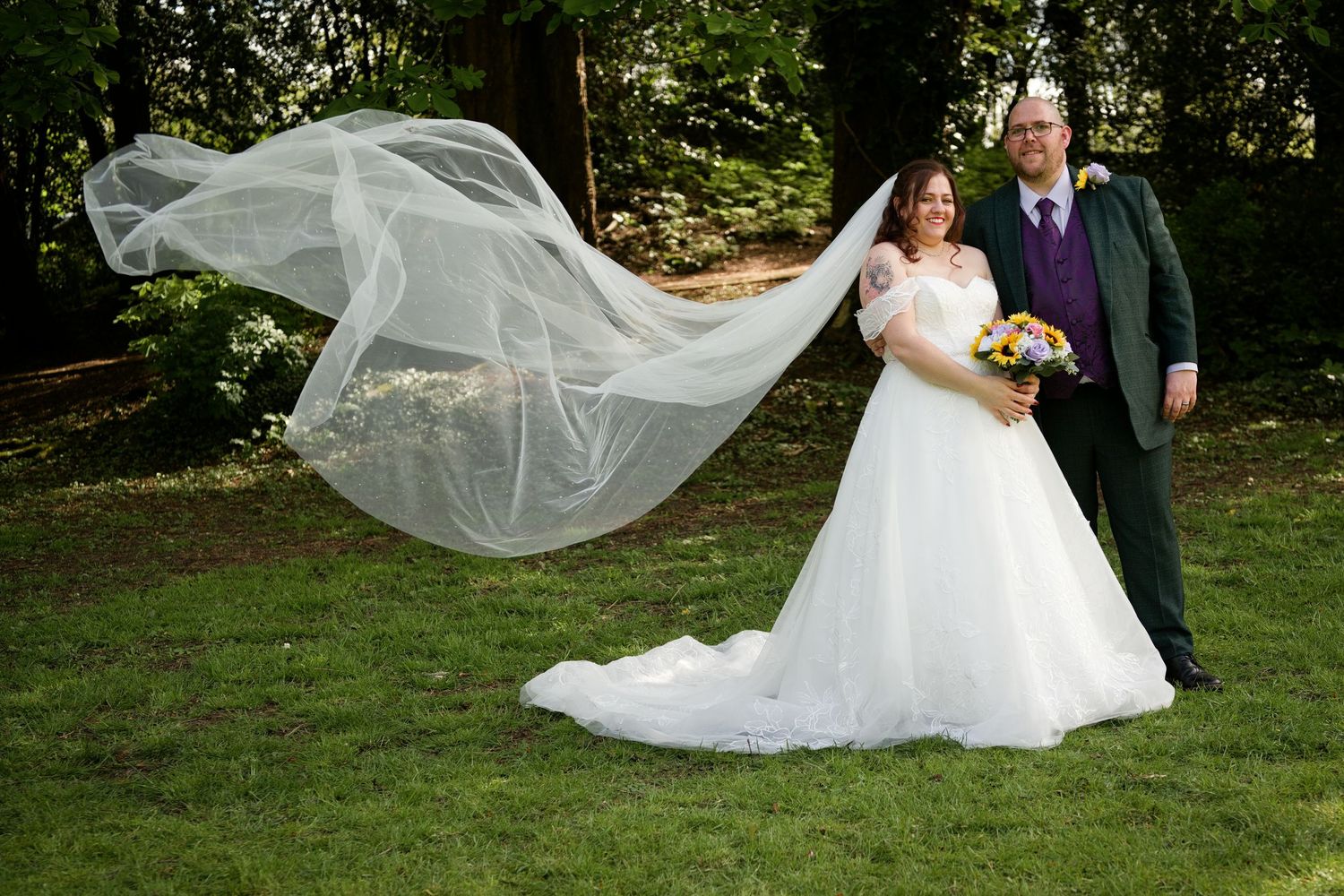 Bride in white ball gown with flowing veil and groom in green suit pose outdoors on grass with trees behind them.