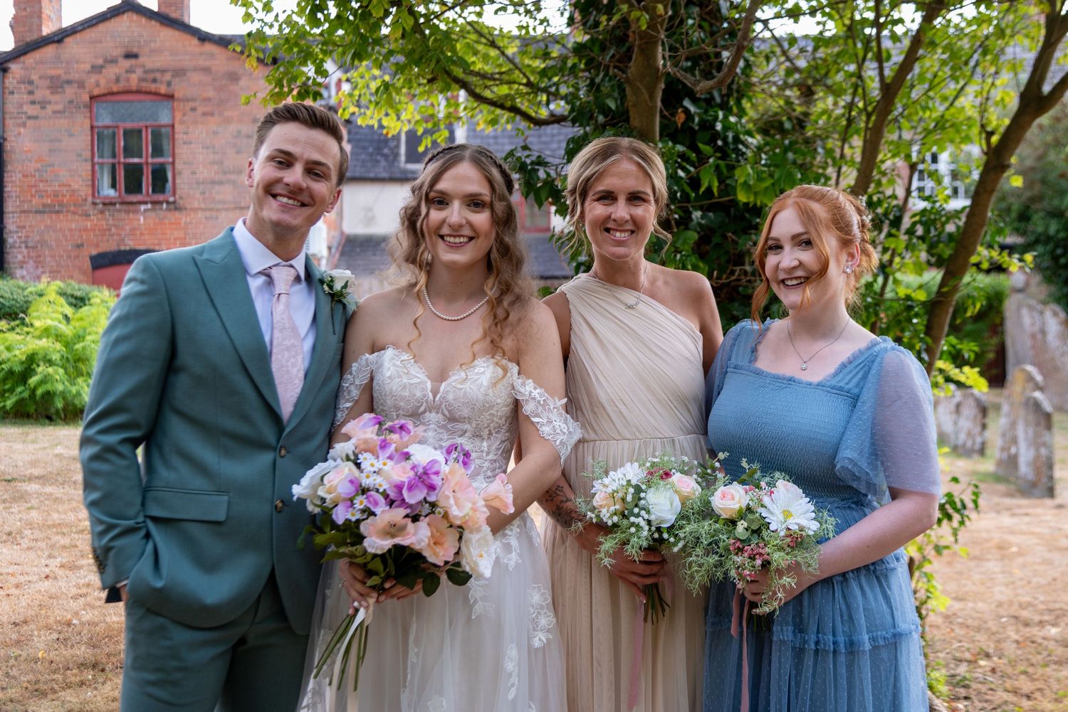 Wedding party posing outdoors, bride in white gown with bouquet flanked by groom in teal suit and two bridesmaids in blue dresses.