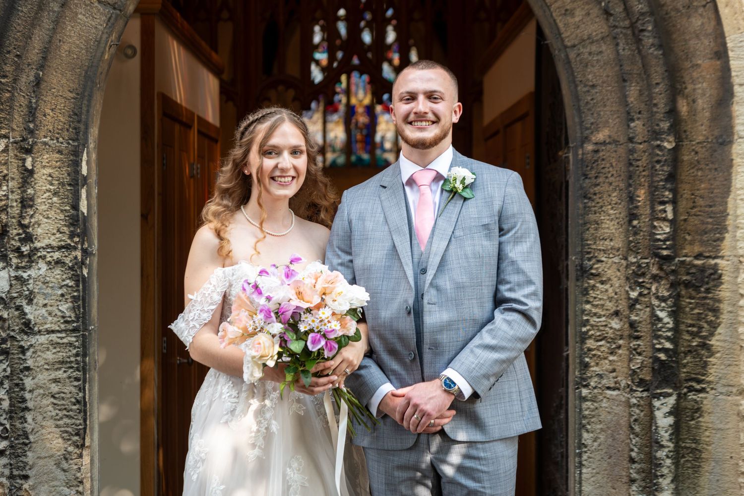 Smiling bride holding colorful bouquet stands with groom in grey suit and pink tie outside church doors.