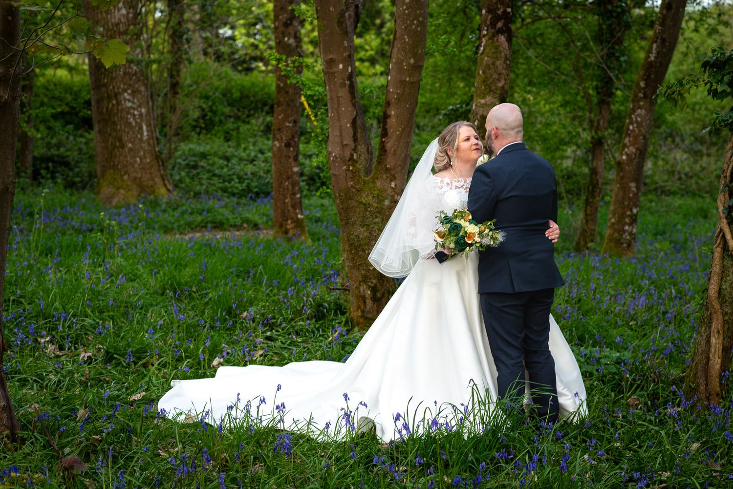A couple embraces in a woodland setting with blooming bluebells carpeting the forest floor, the bride wearing a flowing white gown.