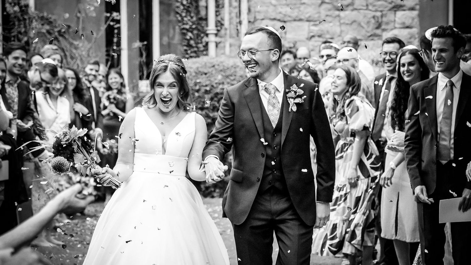 Black and white photo of a joyful bride and groom walking through confetti shower at outdoor wedding ceremony.