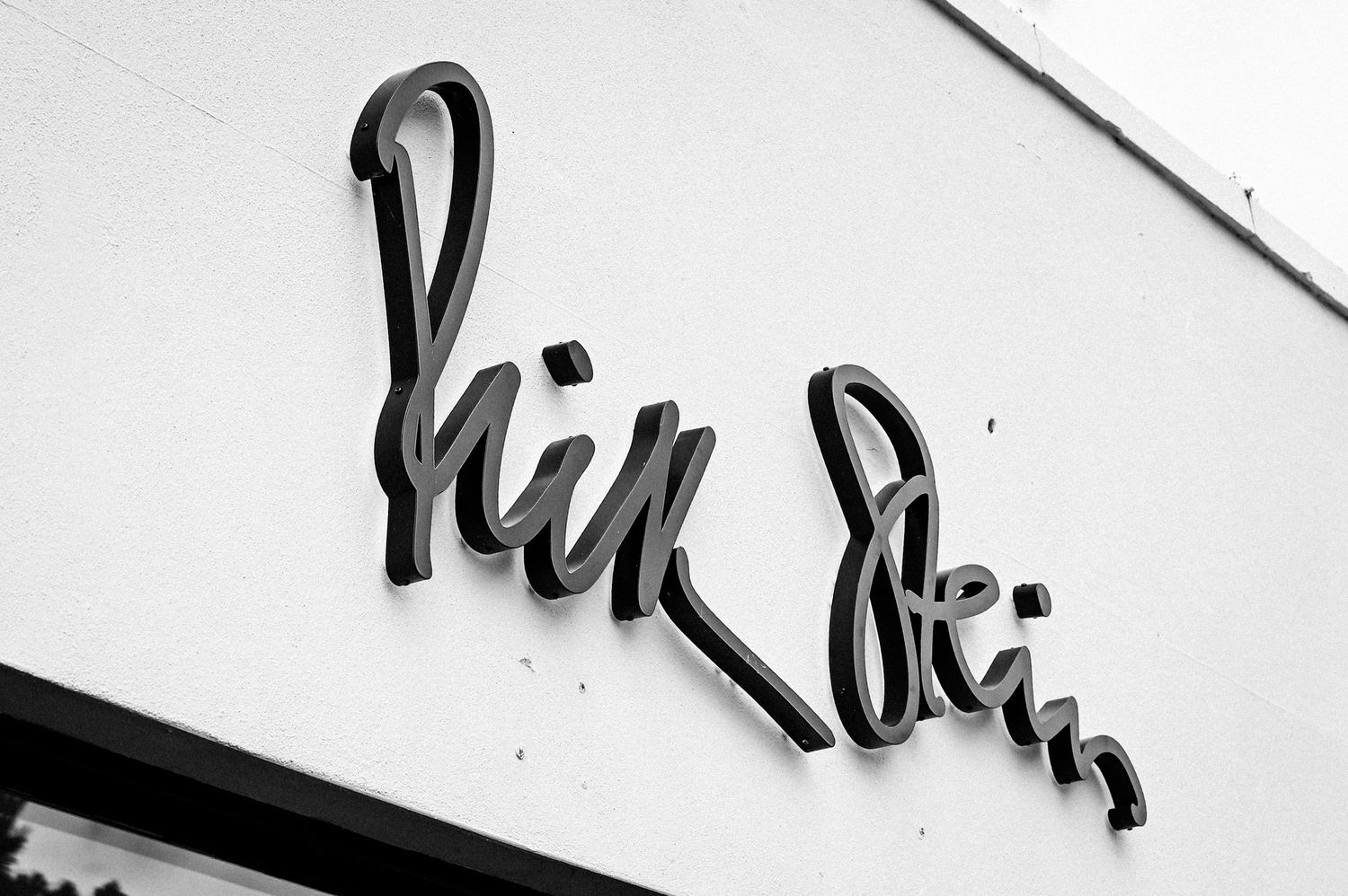 Black and white photo of a cursive script store sign reading Hill Stein mounted on a white wall.