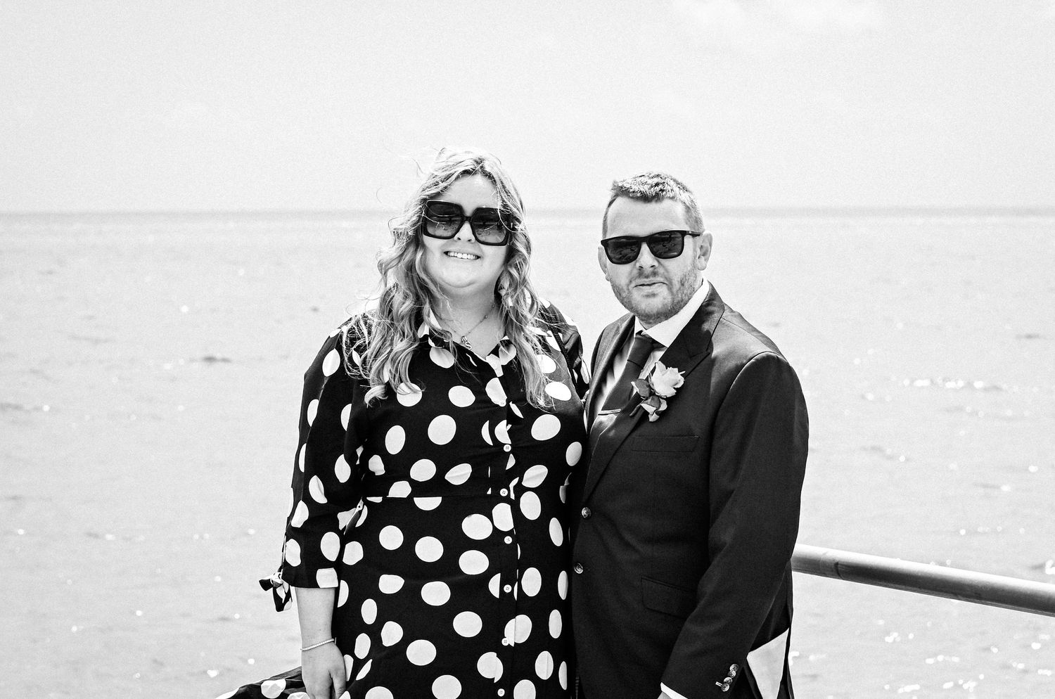 Black and white photo of a smiling couple in sunglasses posing on a beachfront promenade, woman in polka dot dress.