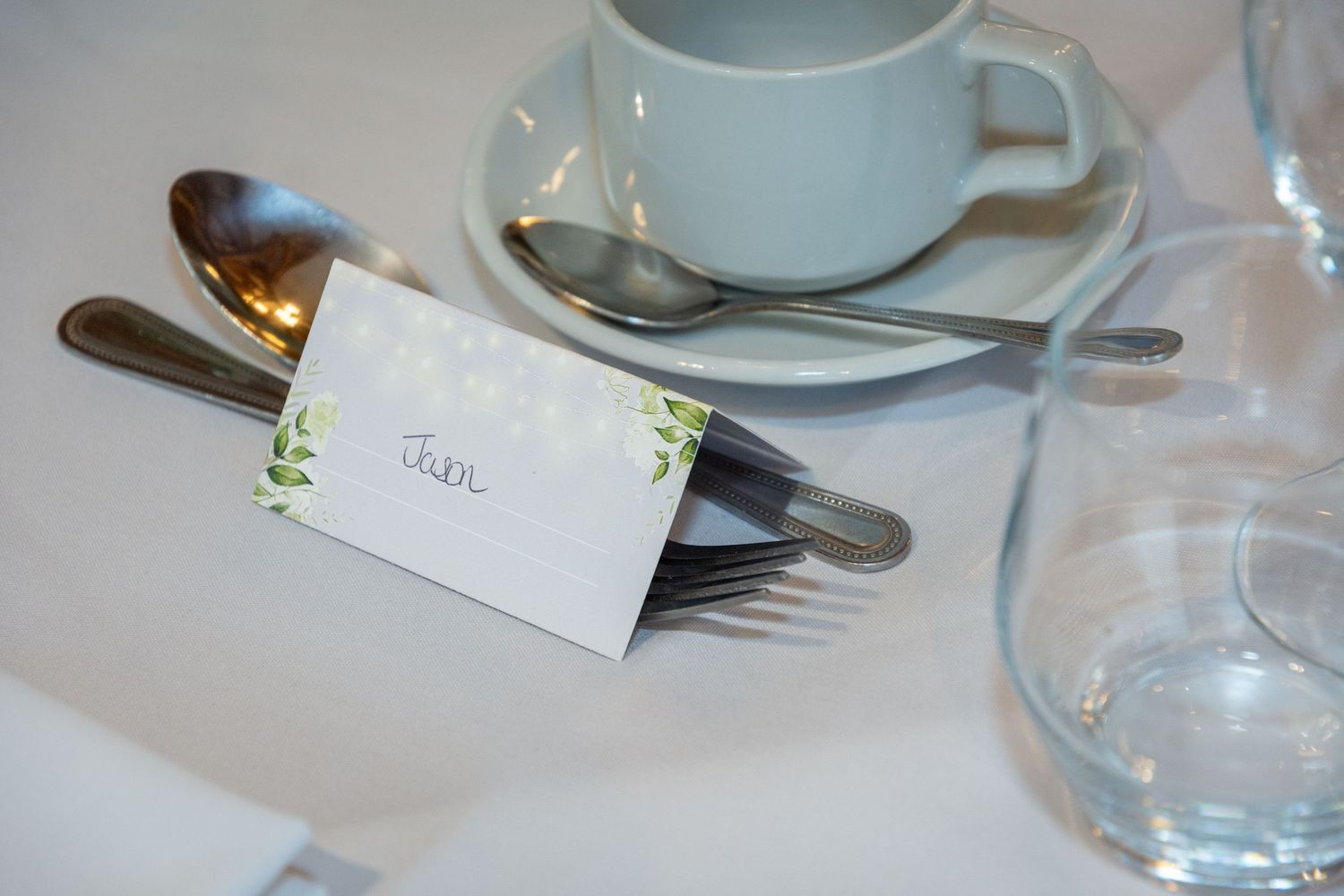 Elegant place setting with a name card, silverware, coffee cup, and glass on a white tablecloth.