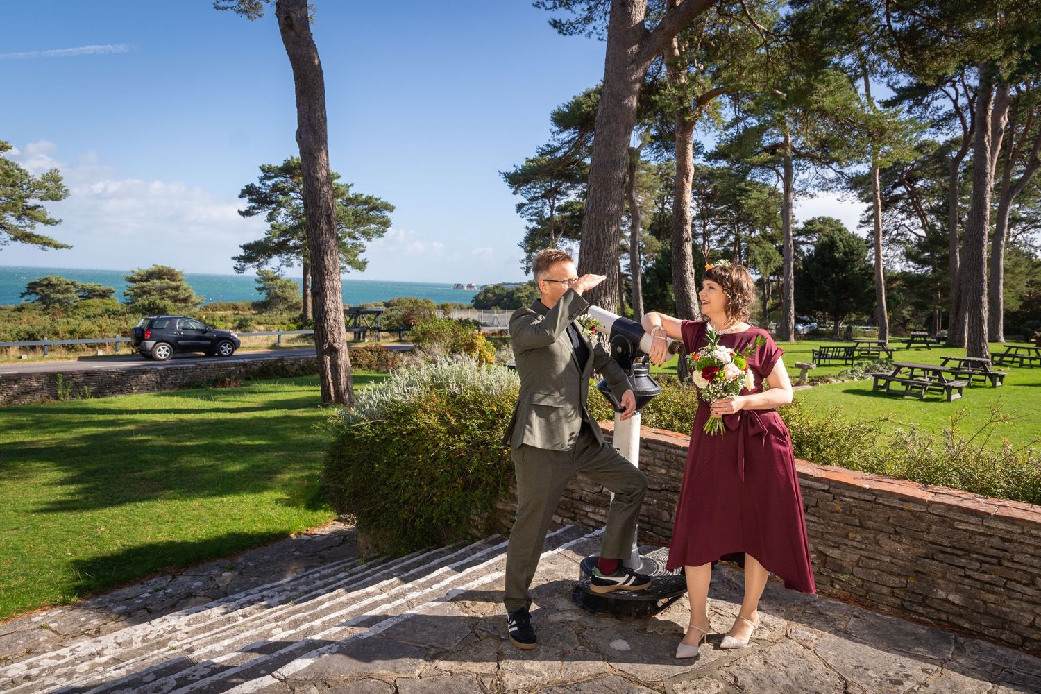 Couple celebrating outdoors at a coastal garden venue, woman in burgundy dress holding flowers, man in suit, ocean view behind.