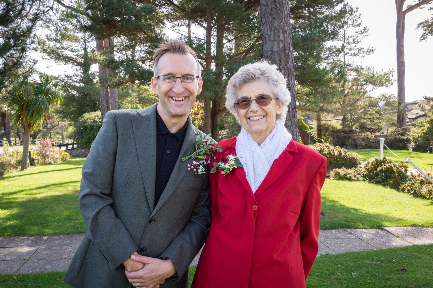 Smiling man in gray suit and elderly woman in red jacket posing together in a garden.