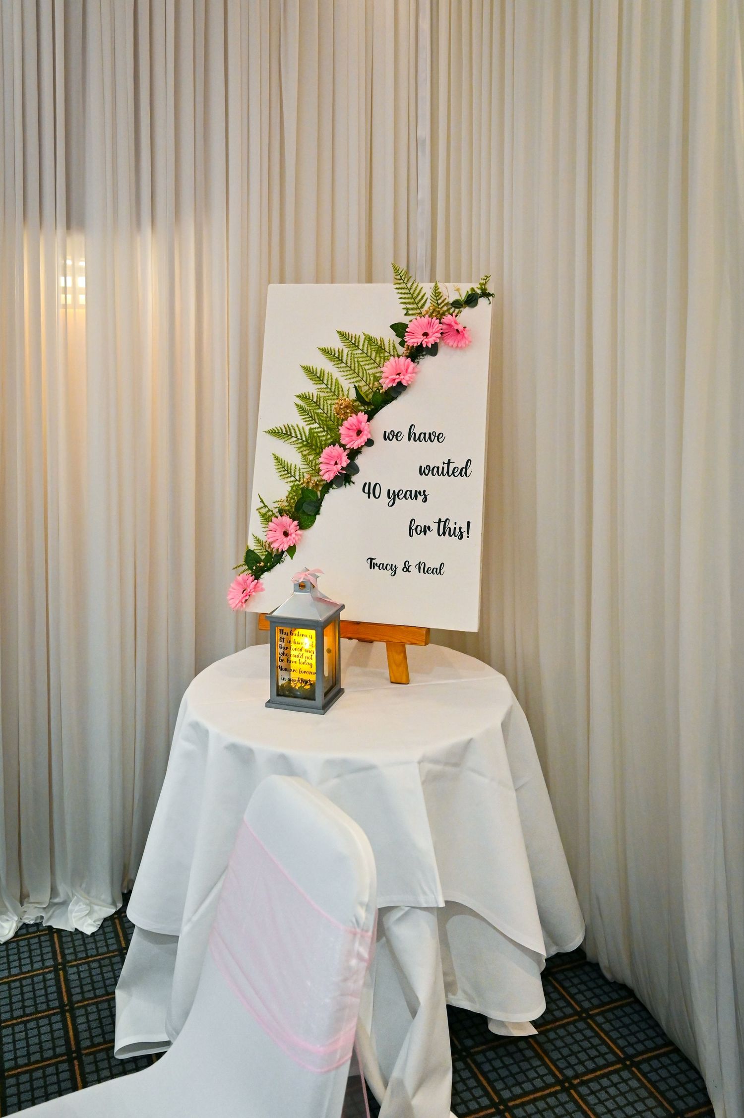Wedding anniversary sign on easel with pink flowers and ferns, reading we have waited 90 years for this on white draped table.