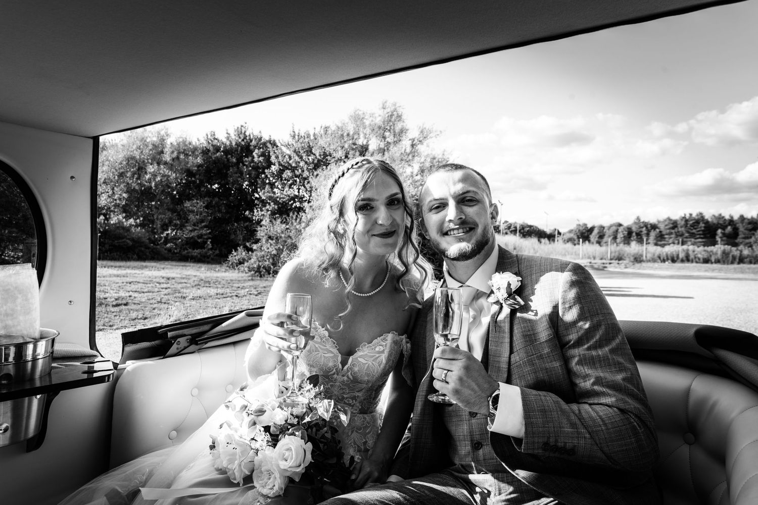 Black and white photo of smiling bride and groom sitting together in a vintage car on their wedding day.