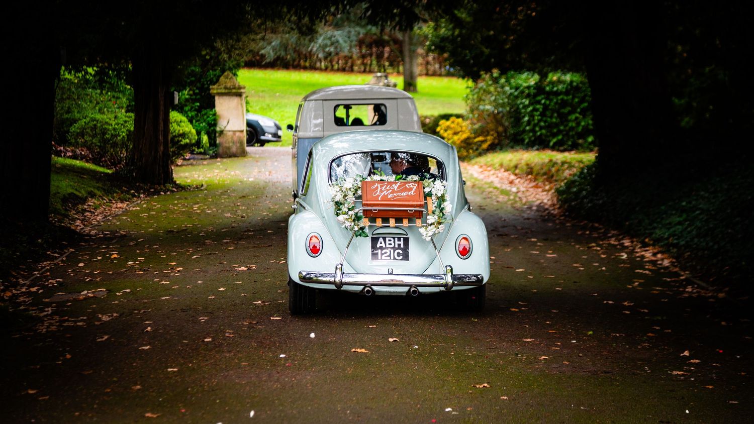 A vintage Volkswagen Beetle drives down a tree-lined path in a park setting.