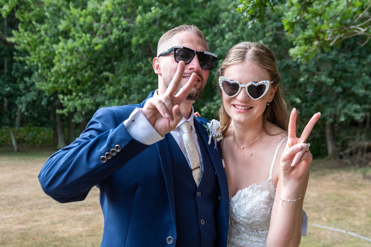 Bride and groom posing outdoors making peace signs, wearing sunglasses, bride in white dress, groom in blue suit.