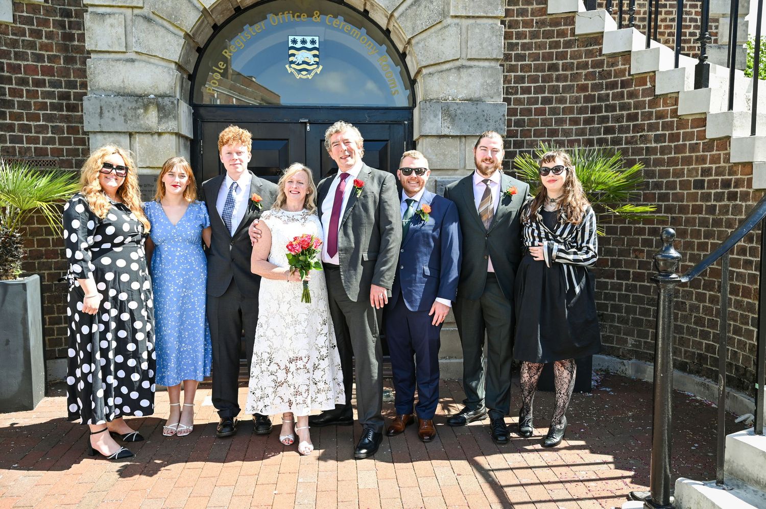 Wedding party poses outside historic brick building, bride in white dress holding bouquet with seven guests in formal attire.