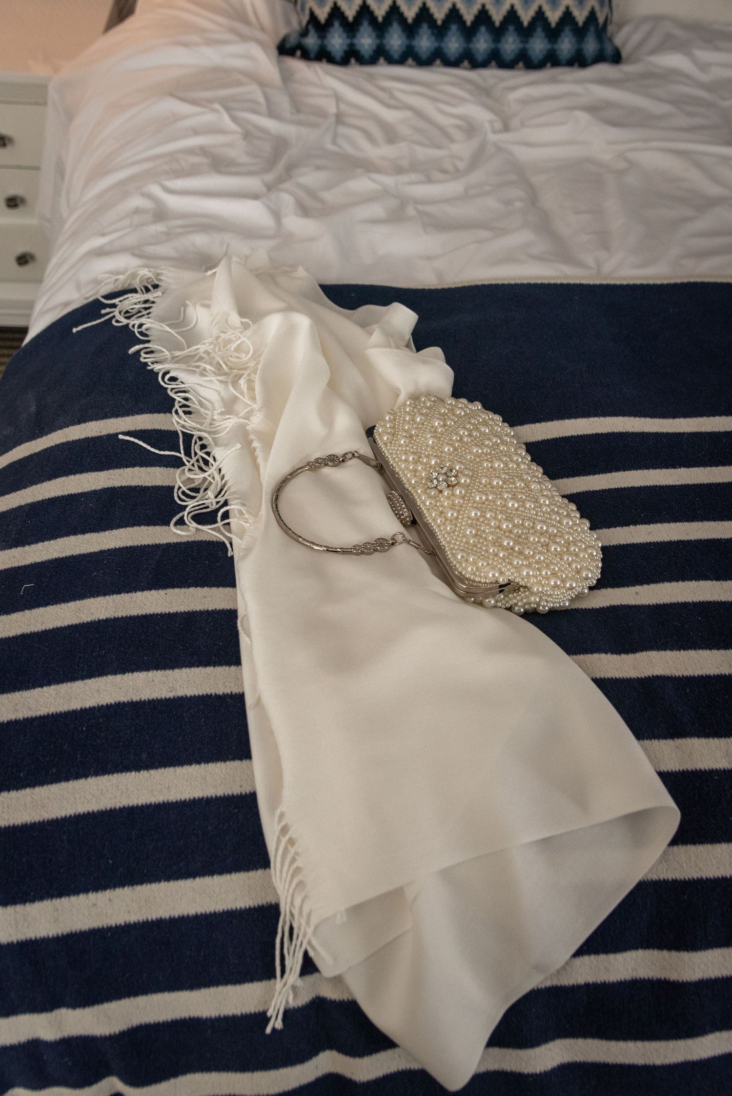 White fringed shawl with pearl beaded clutch and silver bracelet on navy striped bedspread.