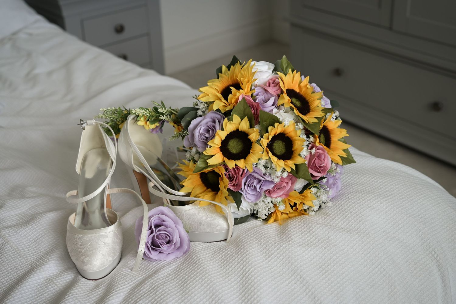 Bridal bouquet with sunflowers and lavender roses beside white heels on a bed.