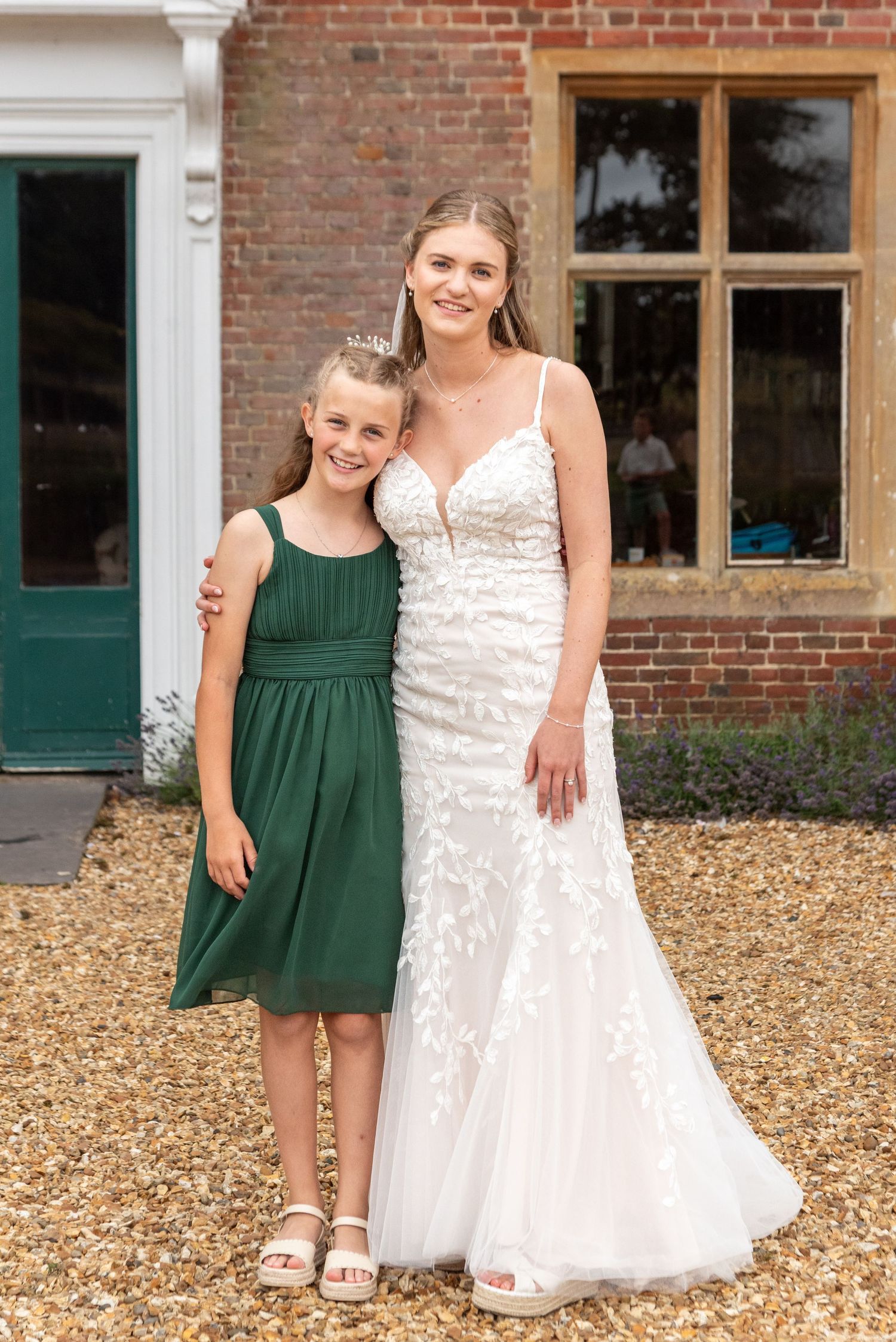 Bride in white lace gown poses with young flower girl in green dress outside brick venue.