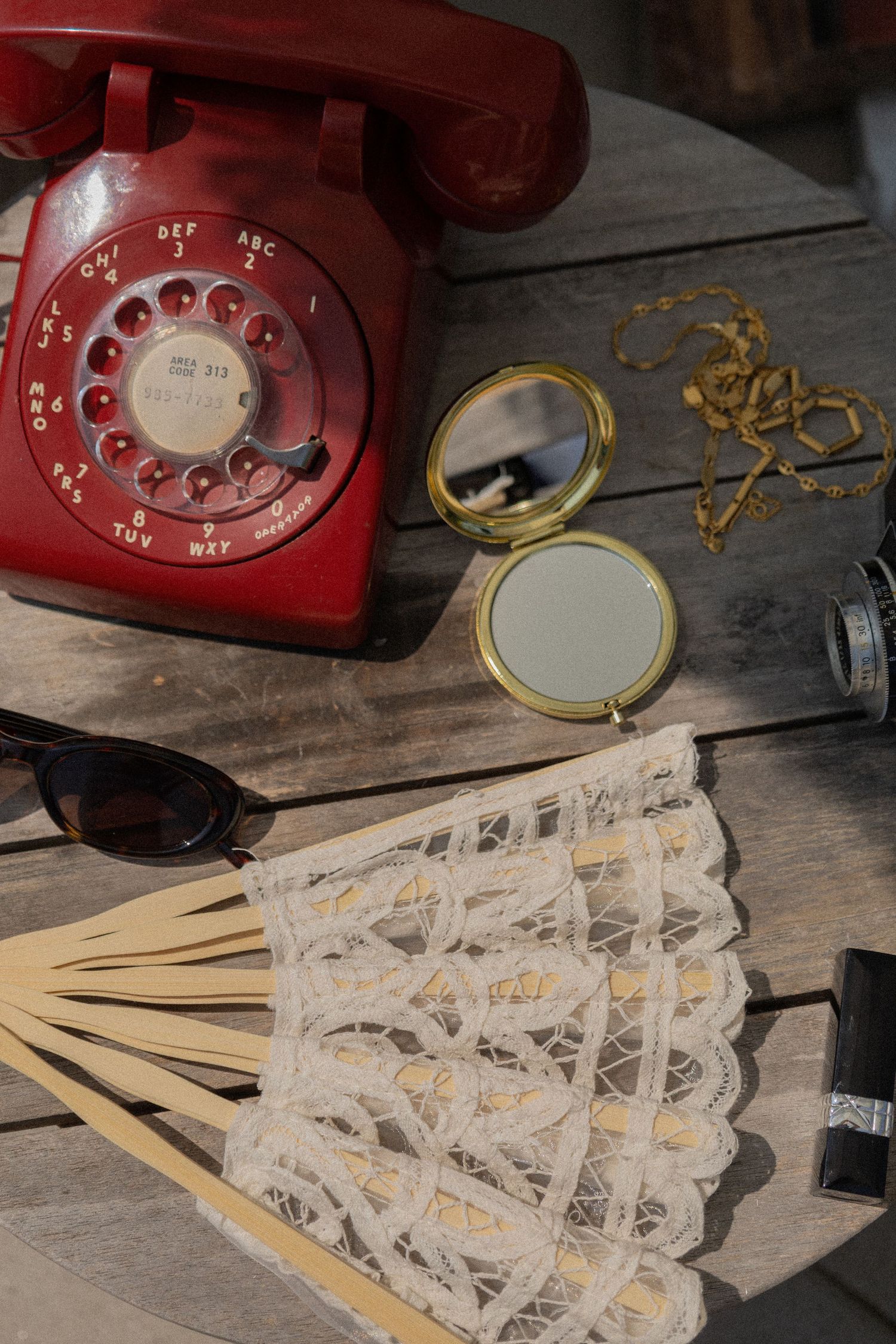 Vintage red rotary phone, lace fan, gold compact mirror, sunglasses, and jewelry arranged on a rustic wooden surface.