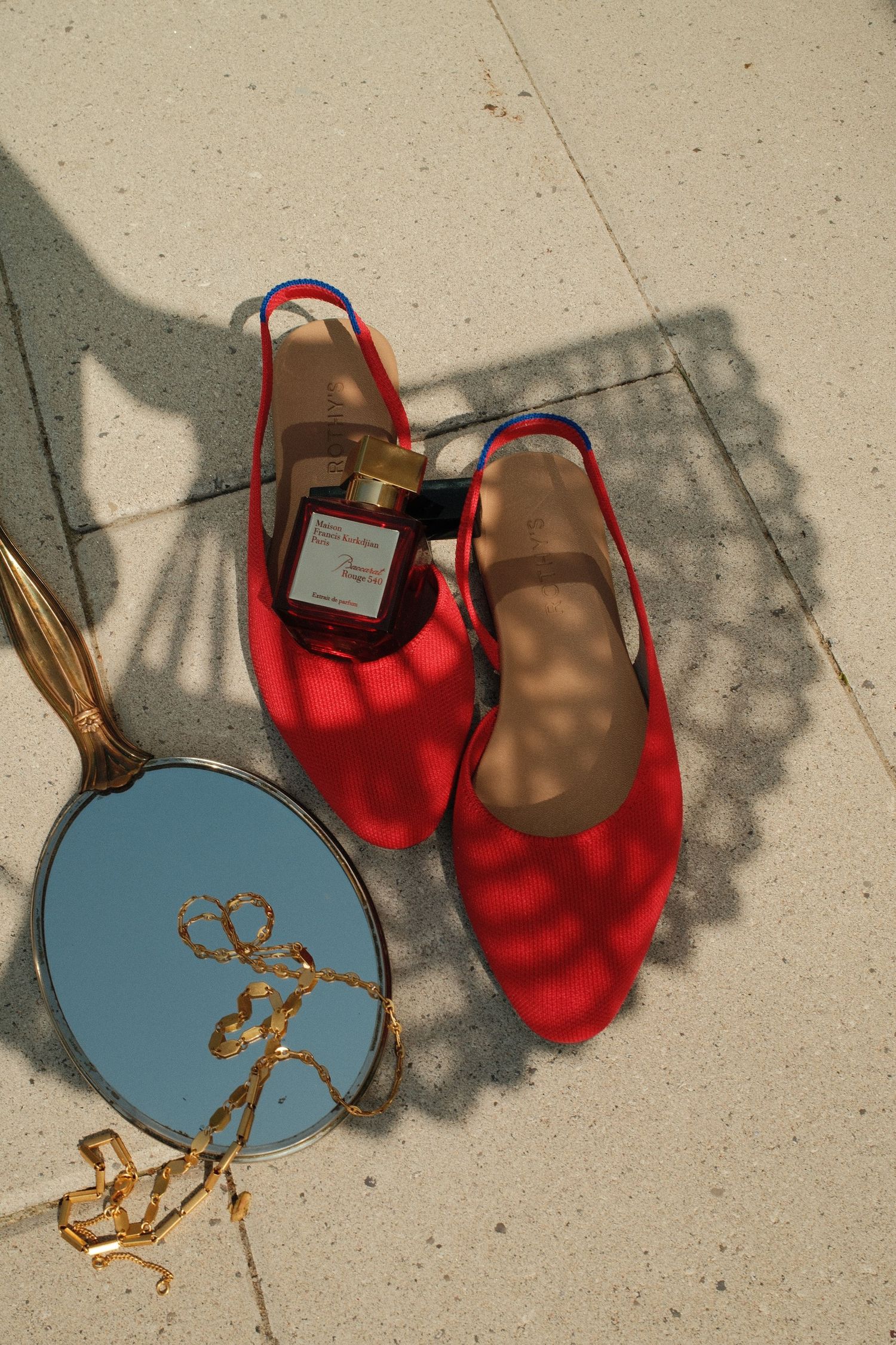 Red slingback heels with perfume bottle and gold jewelry on round mirror, flatlay on beige tile floor.