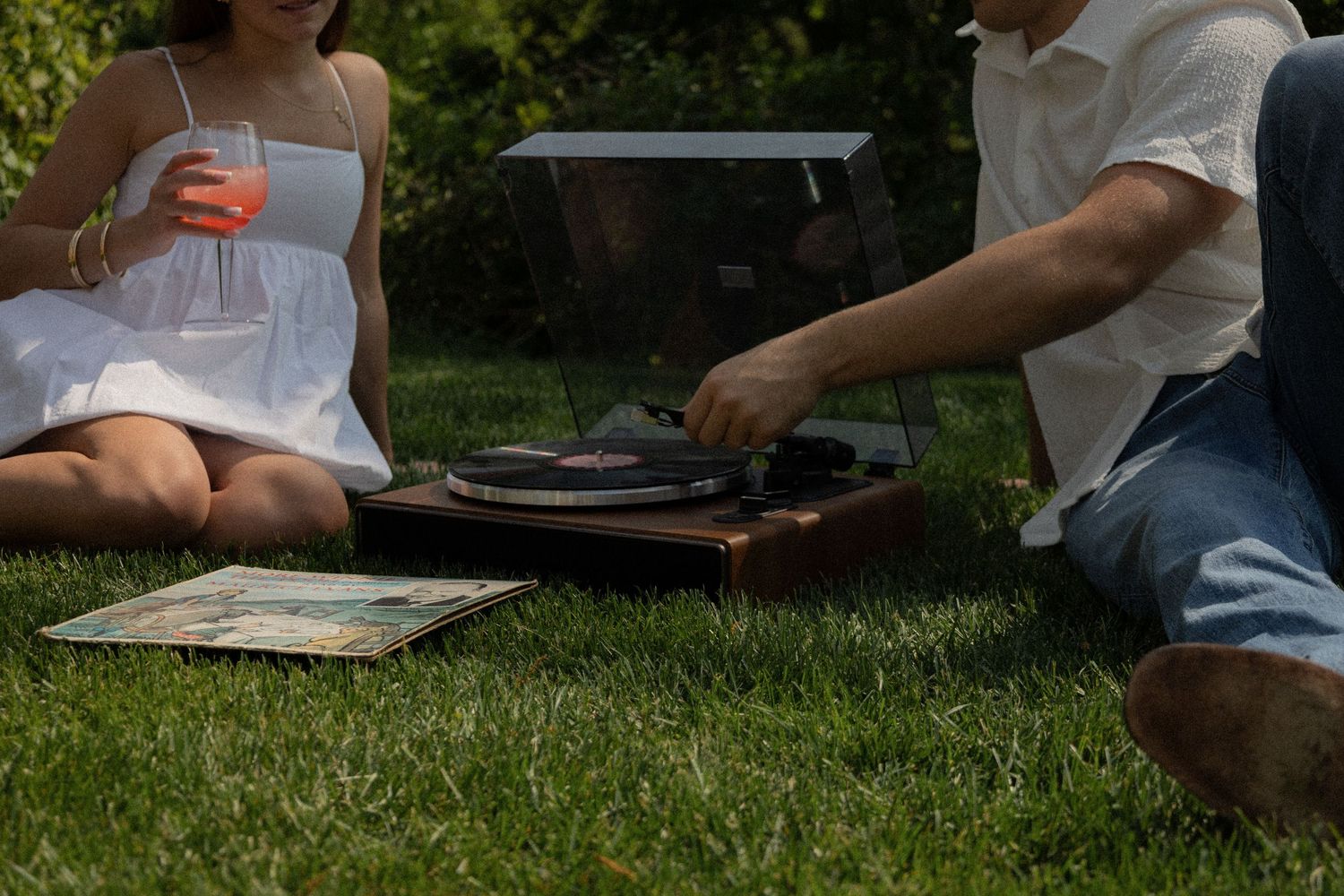 Couple sitting on grass listening to vinyl records on a portable turntable outdoors on a sunny day.