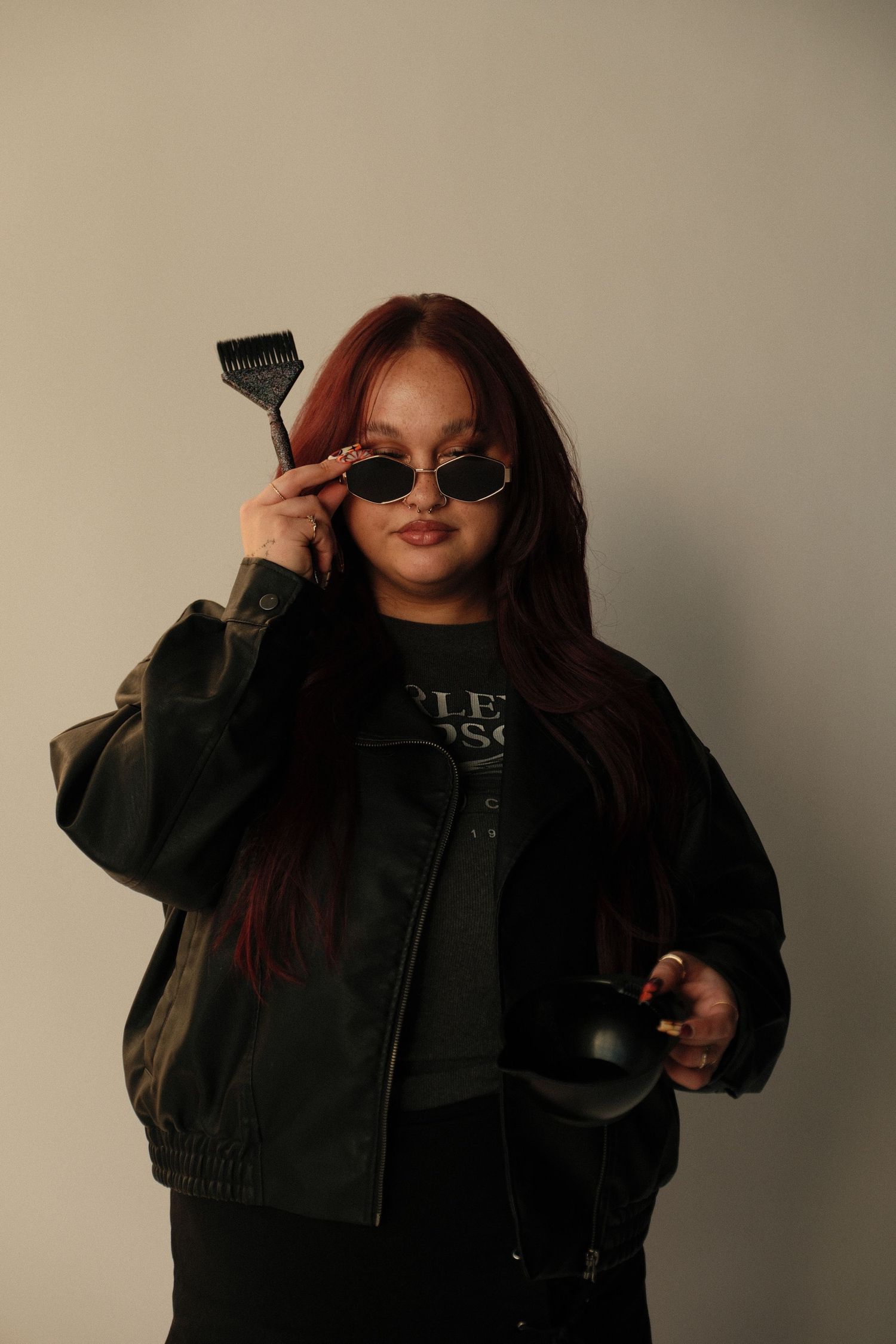 A woman with red hair wearing sunglasses and a dark jacket holds a hair dye brush and bowl against a light background.