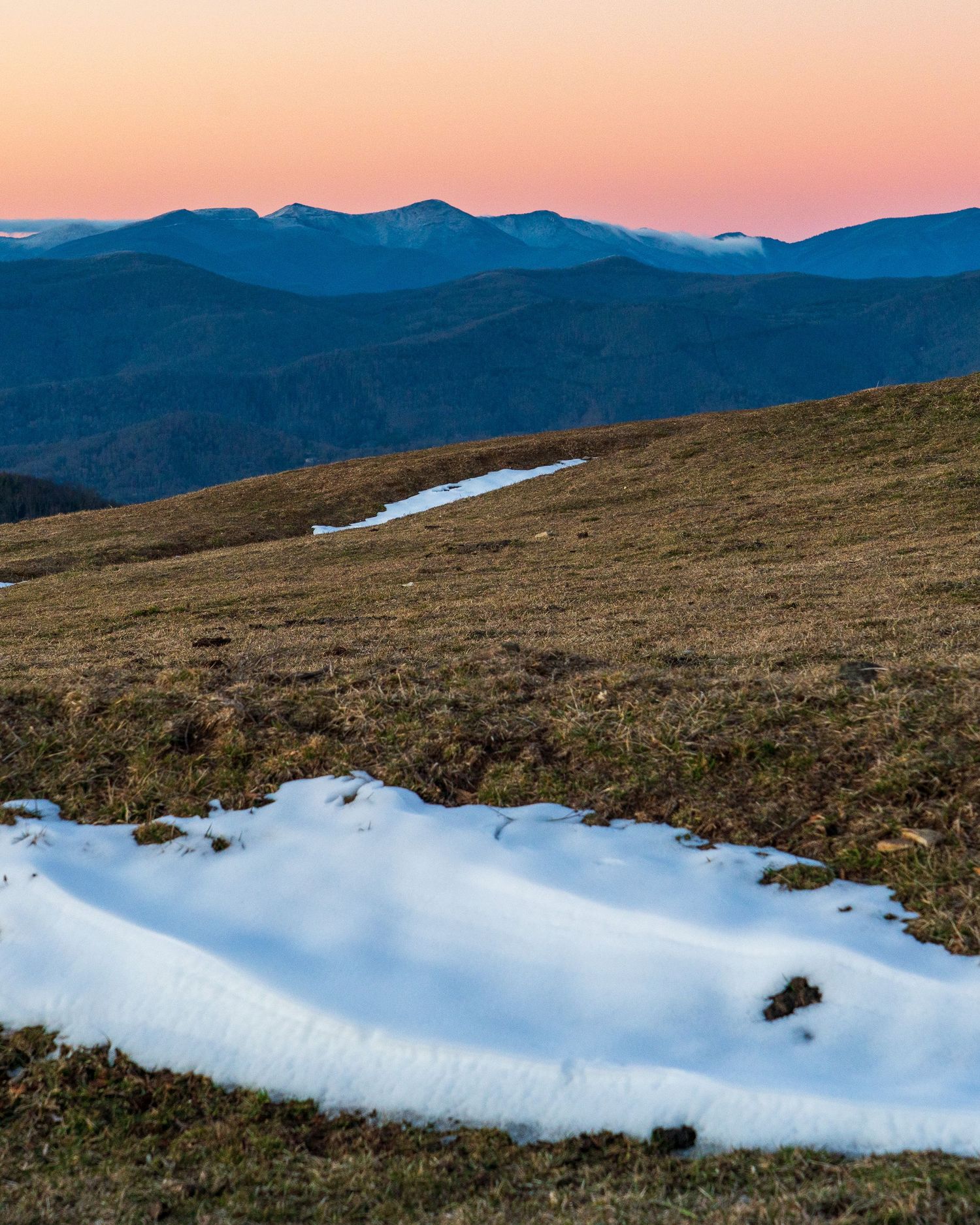 Snow patches on alpine meadow at sunset with blue mountain ridges and pink sky in background.