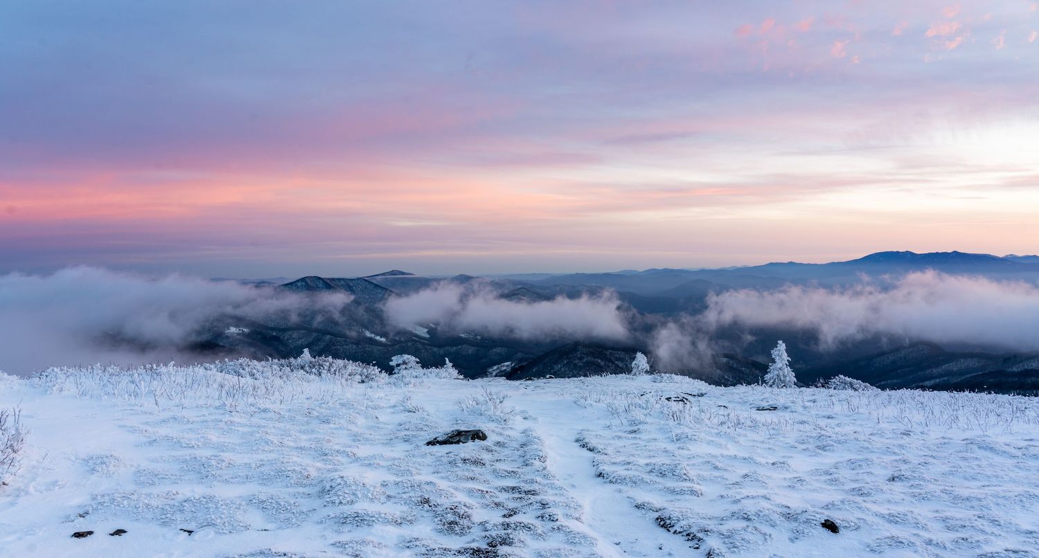 Snow-covered mountain summit at sunset with pink sky, clouds rolling over peaks, and a winding snowy path in winter landscape.
