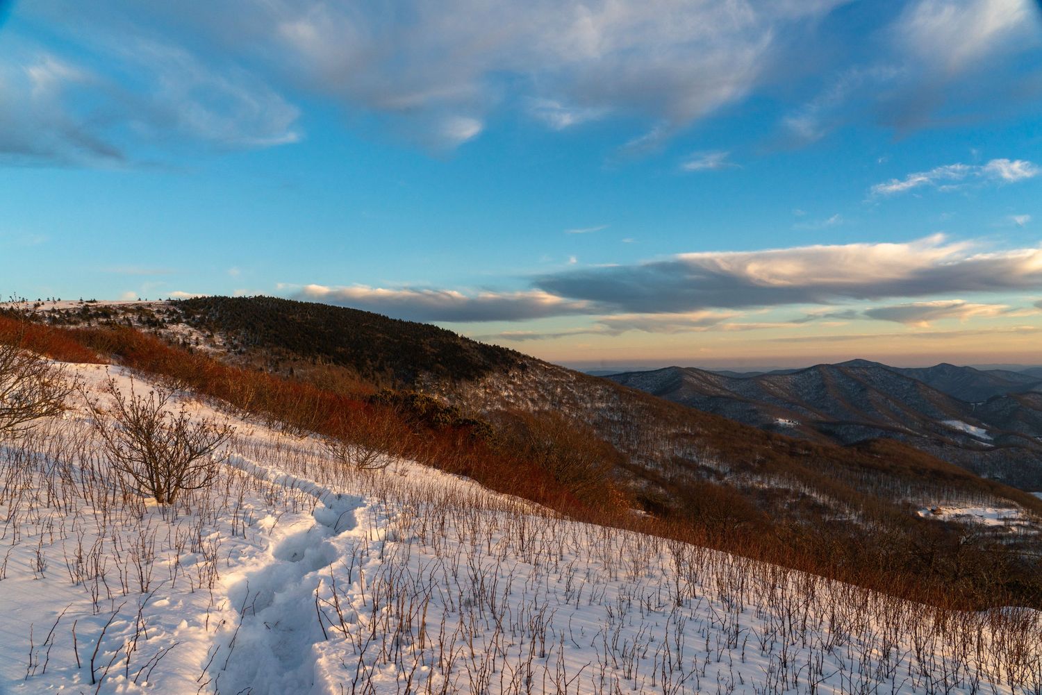 Snow-covered mountain trail winds through winter landscape at golden hour with blue sky and distant peaks.
