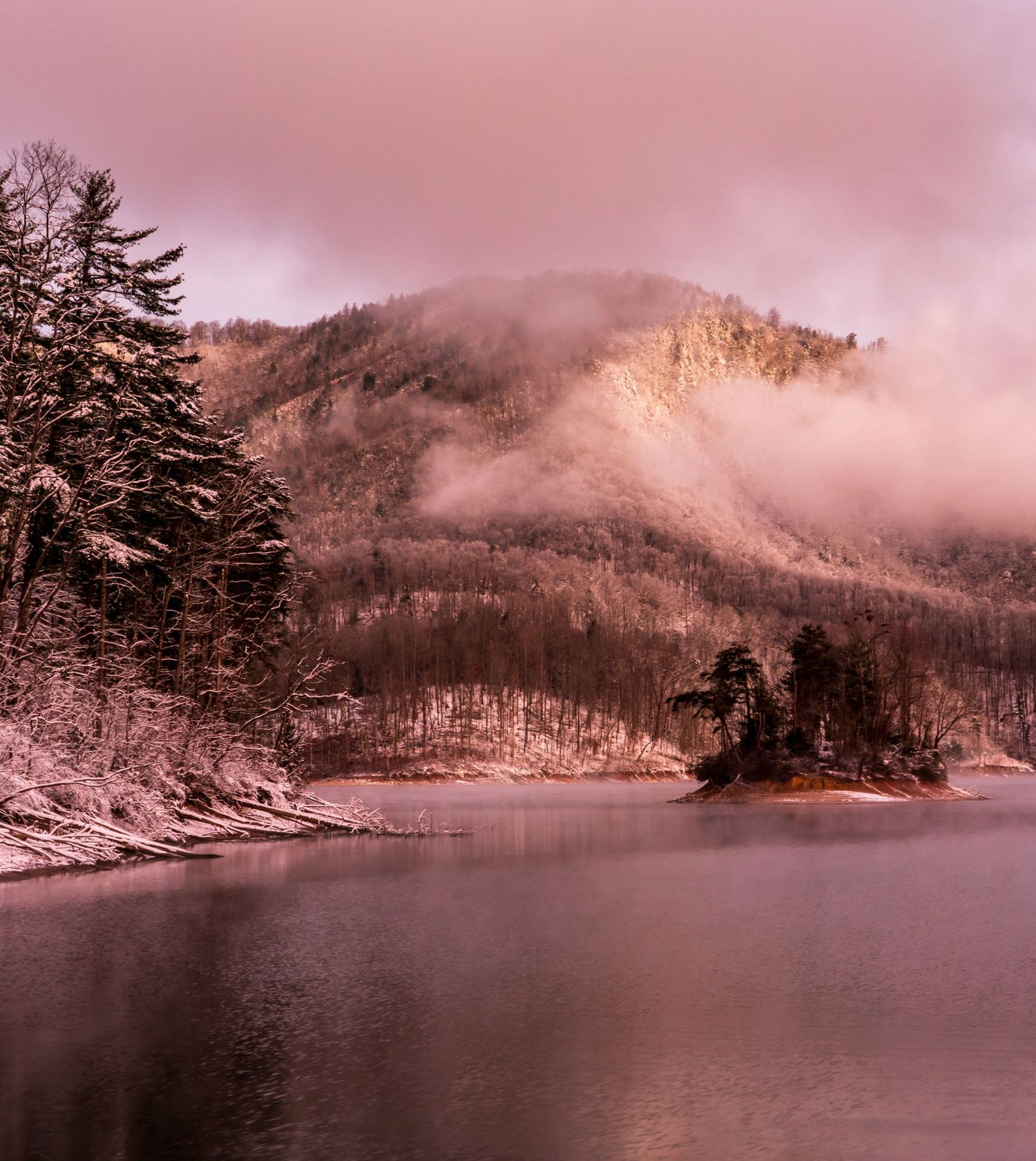 Snow-dusted mountain reflected in calm lake waters under a pink misty sky in winter.