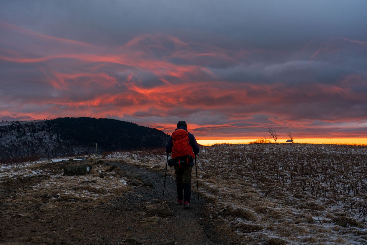 Hiker with red backpack and trekking poles walks a snowy trail under a dramatic fiery sunset sky.