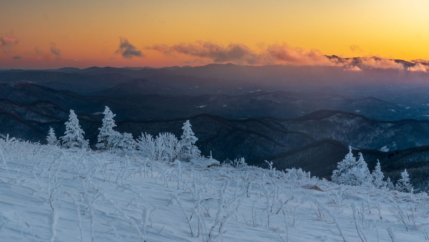 Snow-covered mountain slope at sunset with frost-laden trees and colorful orange sky over misty blue ridgelines.