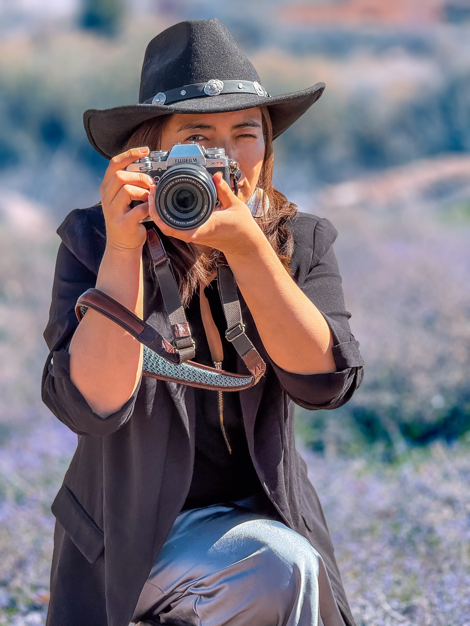 Woman in black hat and blazer crouching outdoors, holding a DSLR camera up to her eye while photographing.