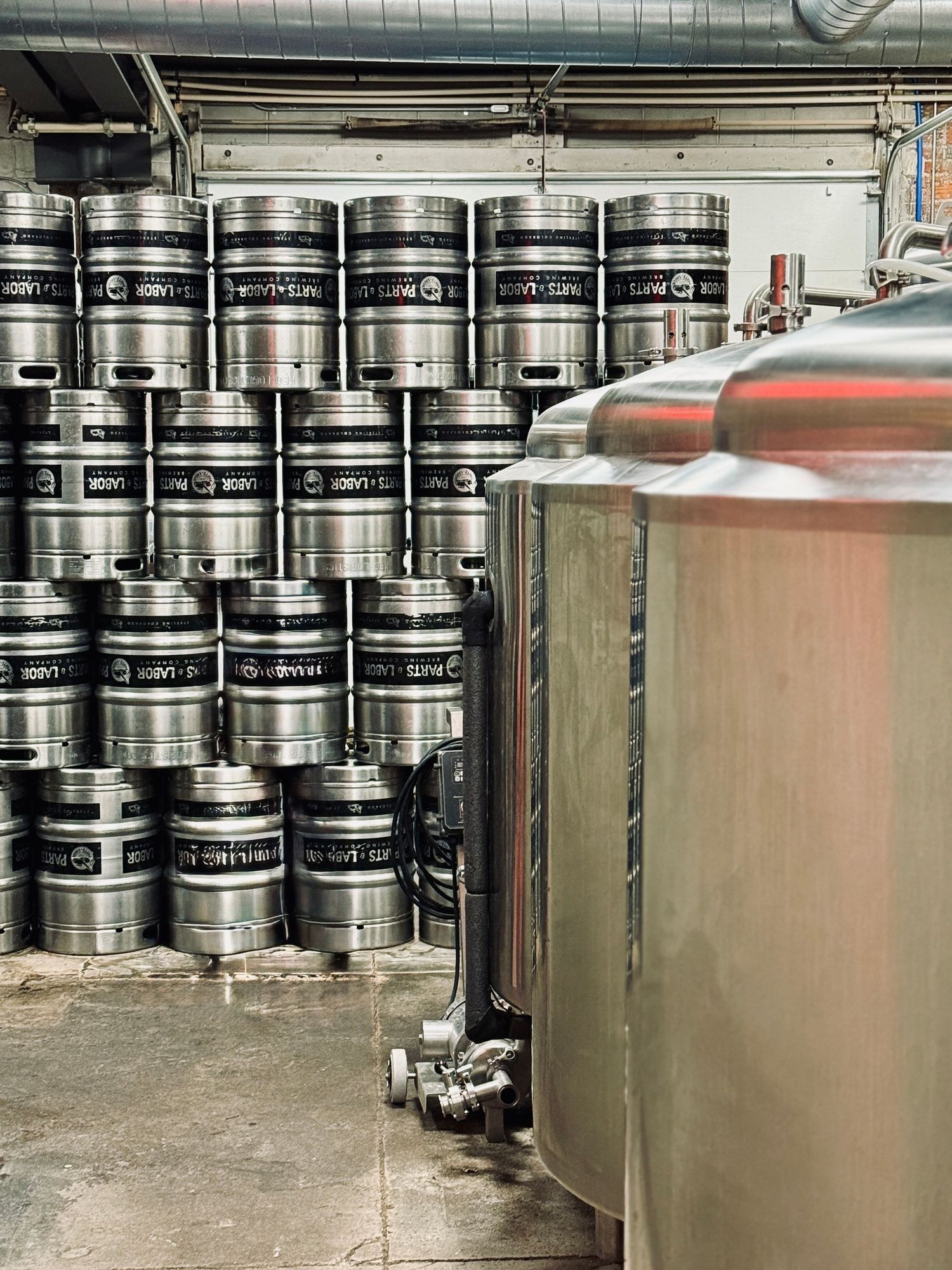 Stacked metal kegs and a large stainless steel brewing tank inside a brewery production facility.