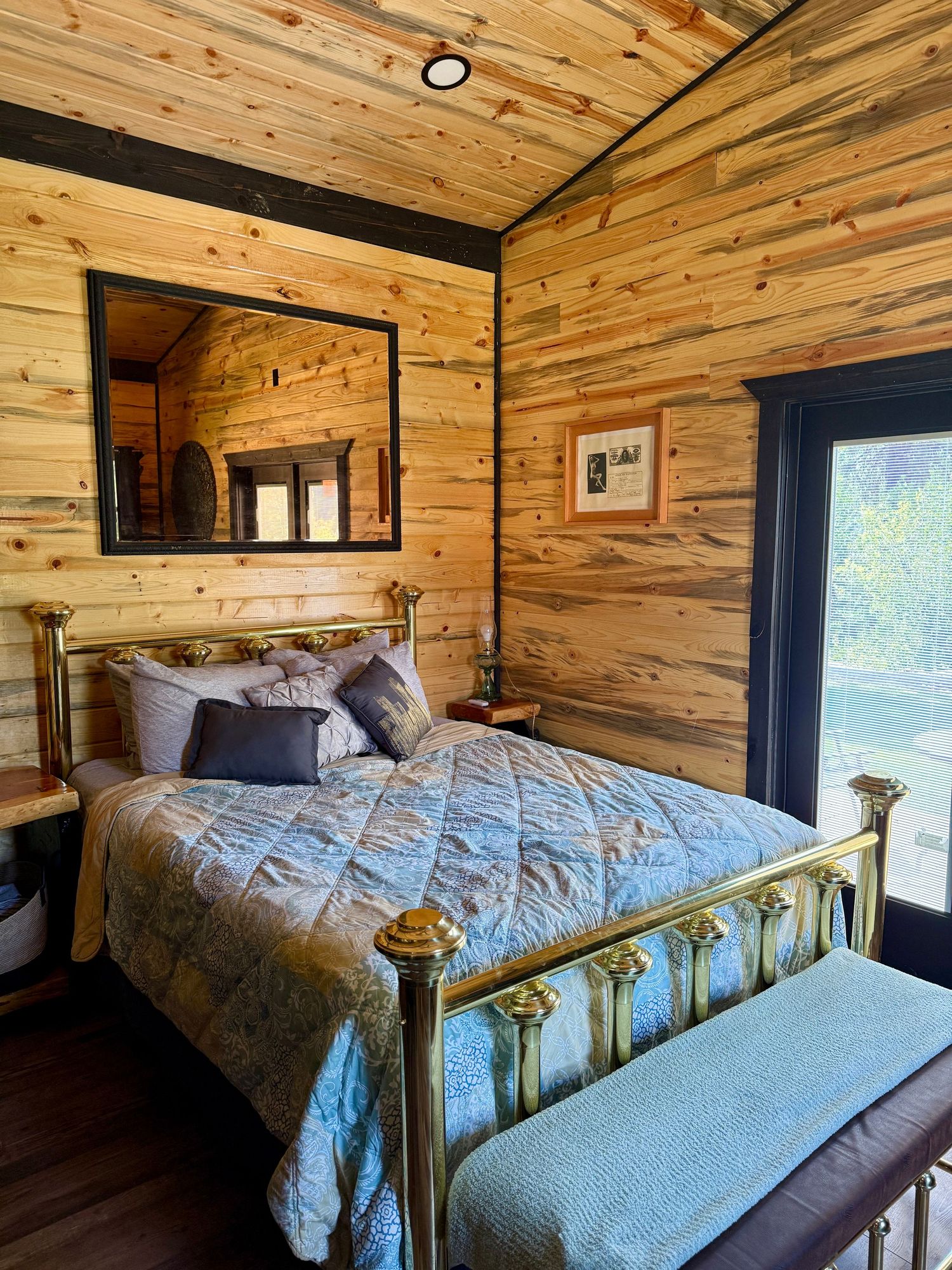 Rustic cabin bedroom with knotty pine wood walls, brass bed frame, and sliding glass door to nature view.