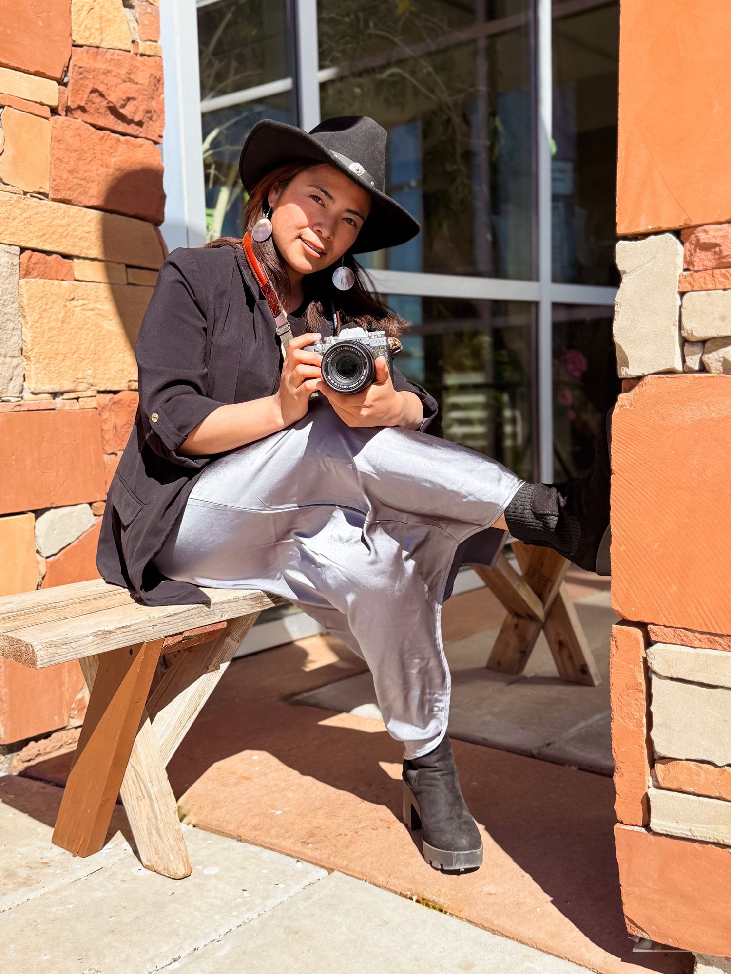 Person in black hat and shirt holding a camera, sitting on a wooden bench outdoors near a brick building.