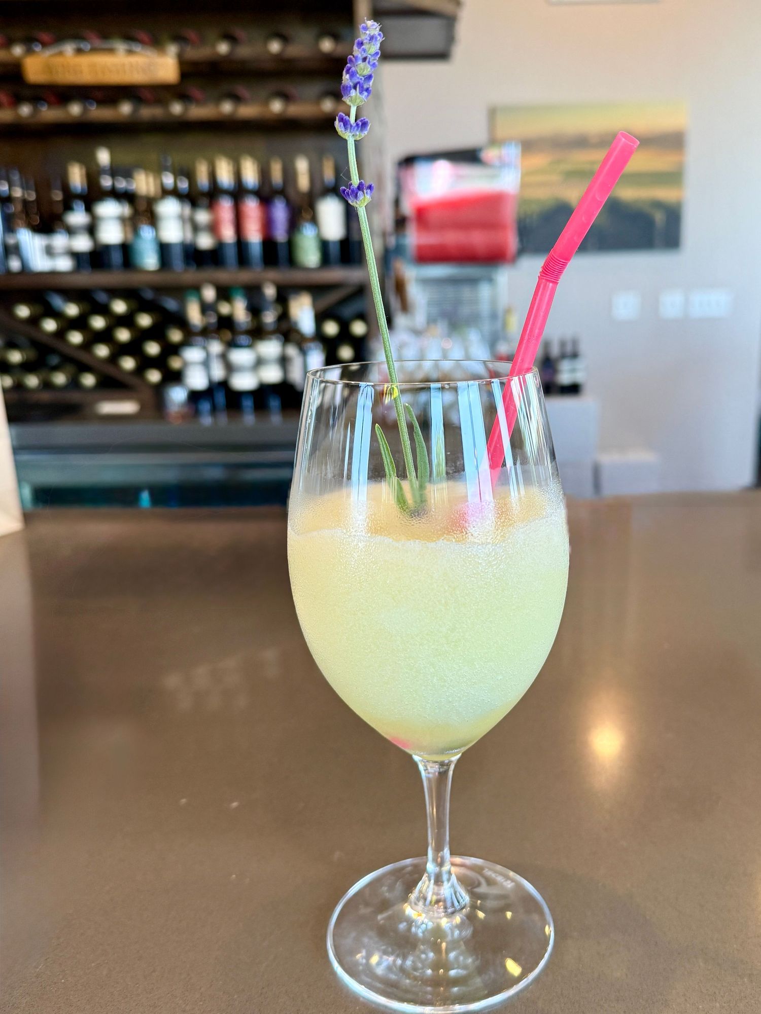 A lavender-garnished pale yellow cocktail in a wine glass at a bar with wine bottles in background.