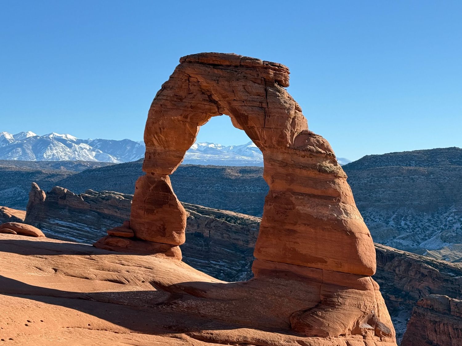 Delicate Arch natural sandstone formation at Arches National Park with snow-capped La Sal Mountains in background.