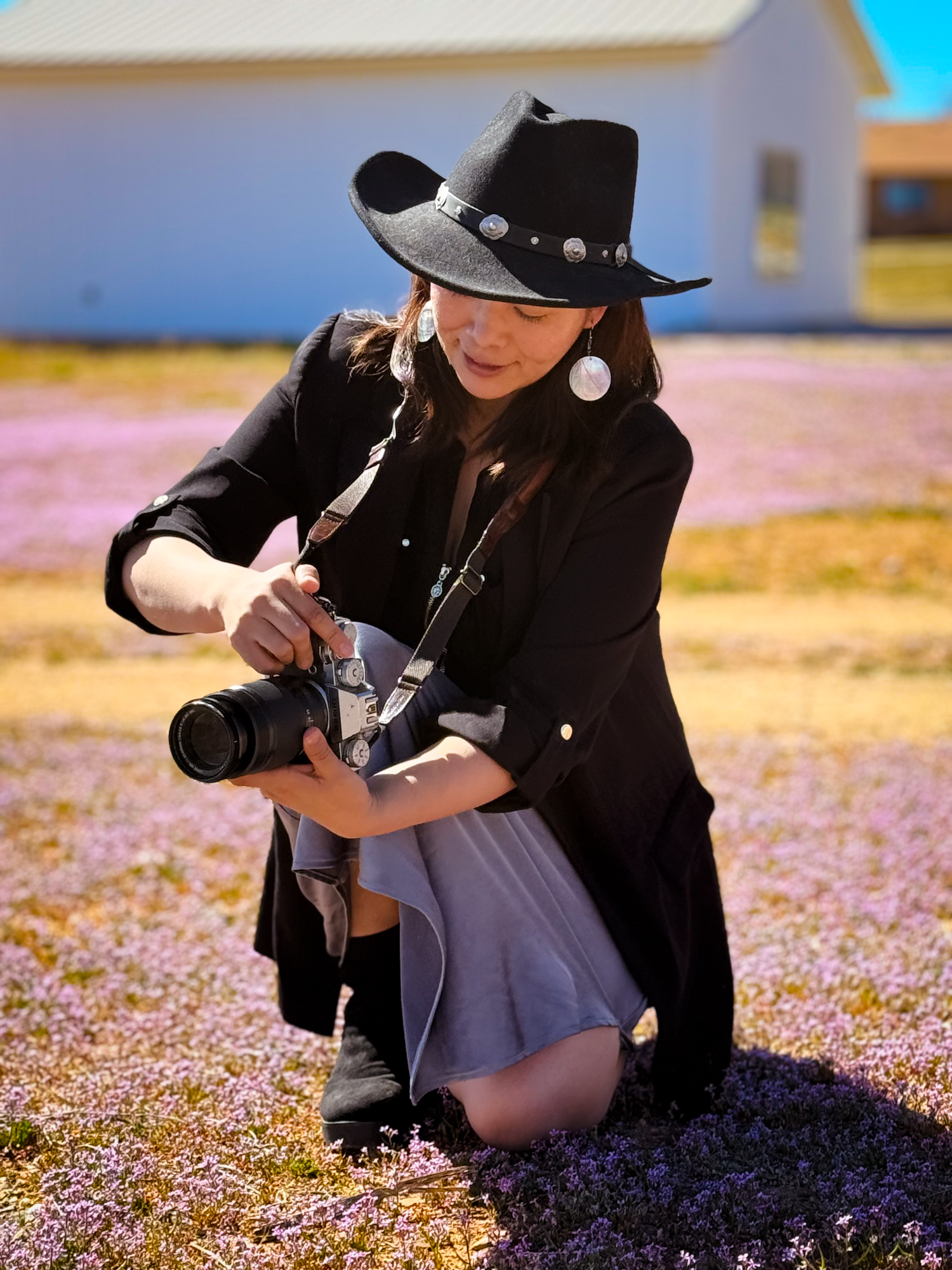 Woman in black cowboy hat crouches in purple wildflower field, adjusting camera settings outdoors.