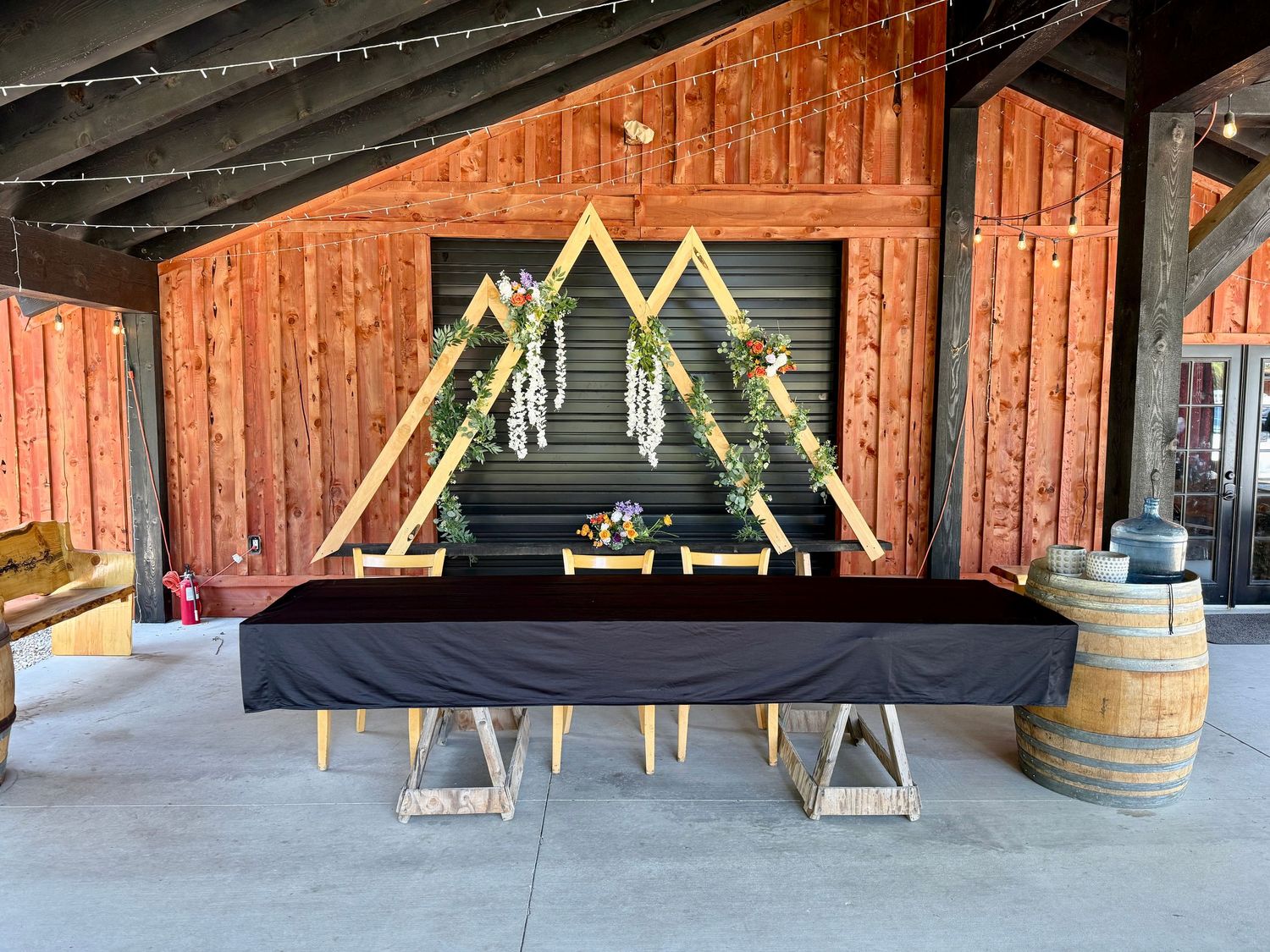 Rustic barn wedding venue with geometric wooden arch backdrop, black draped table, and string lights.