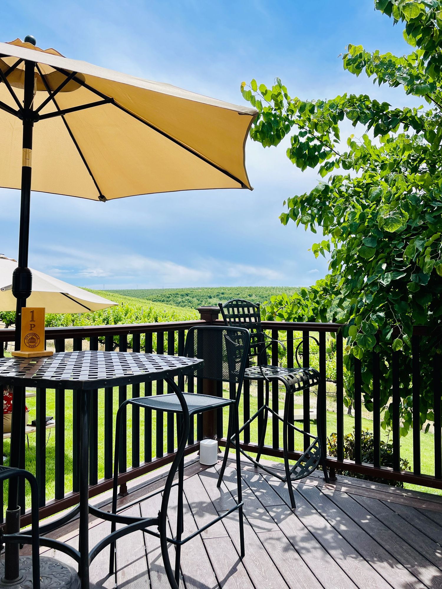 Outdoor deck with black metal bistro table, chairs, and yellow umbrella overlooking lush green vineyard hills.