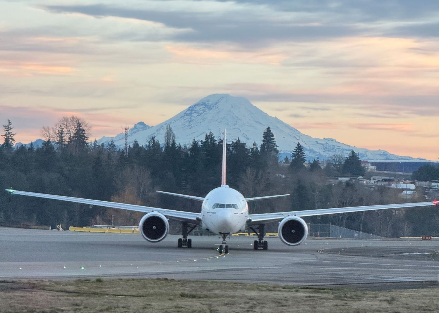 White commercial airplane taxiing on runway with snow-capped Mount Rainier and colorful sunset sky in background.