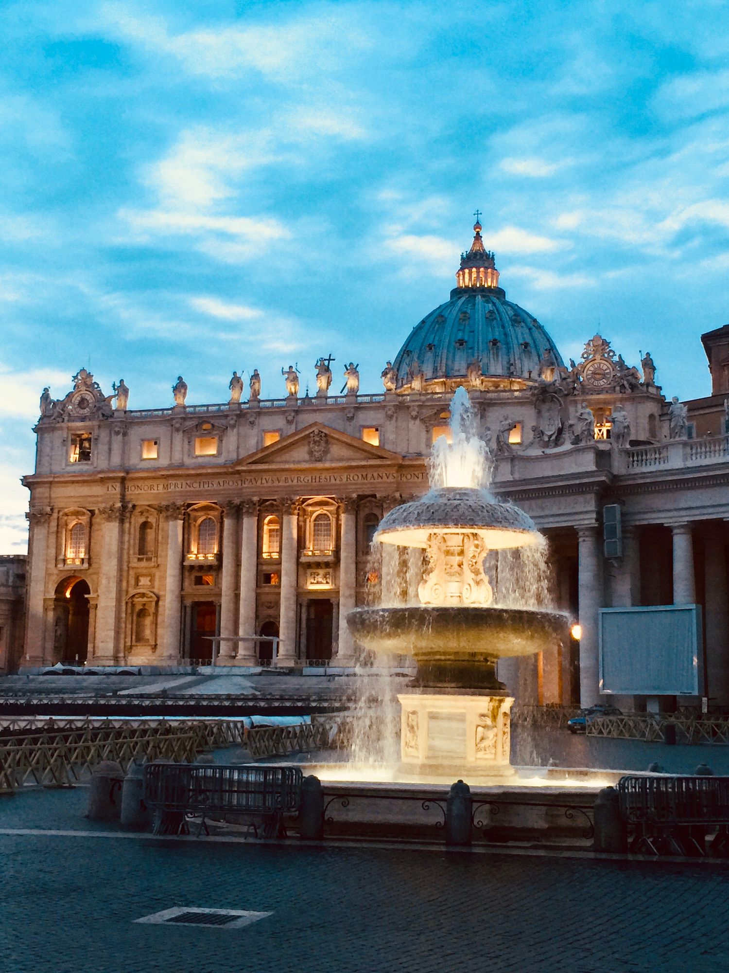 St. Peter's Basilica illuminated at dusk with a glowing fountain in St. Peter's Square, Vatican City.