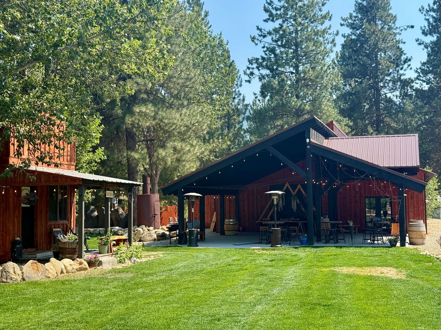 Rustic red barn venue with string lights, outdoor seating, and tall pine trees on green lawn.