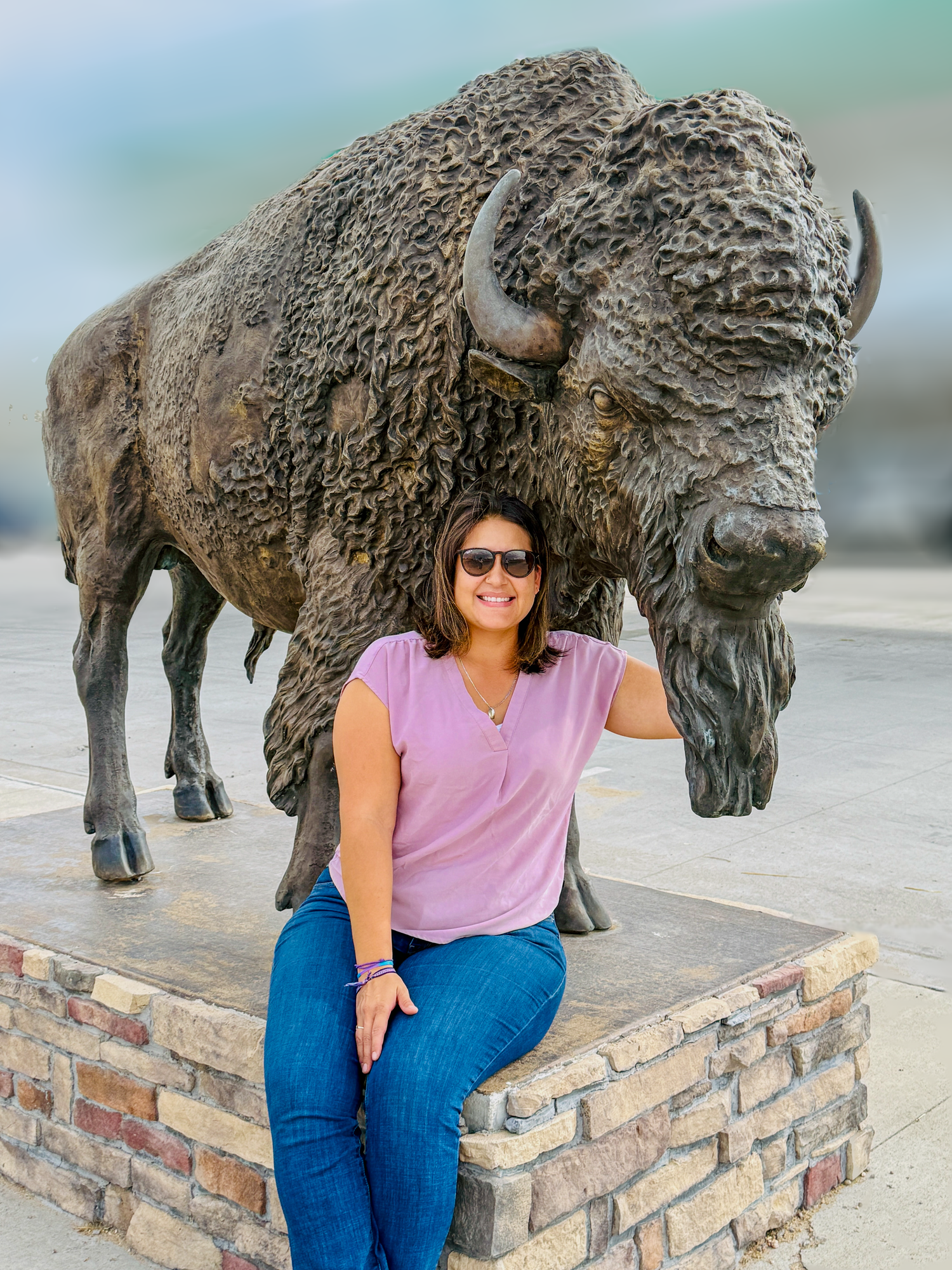 Woman in pink top sitting on base of large bronze bison sculpture at outdoor landmark.
