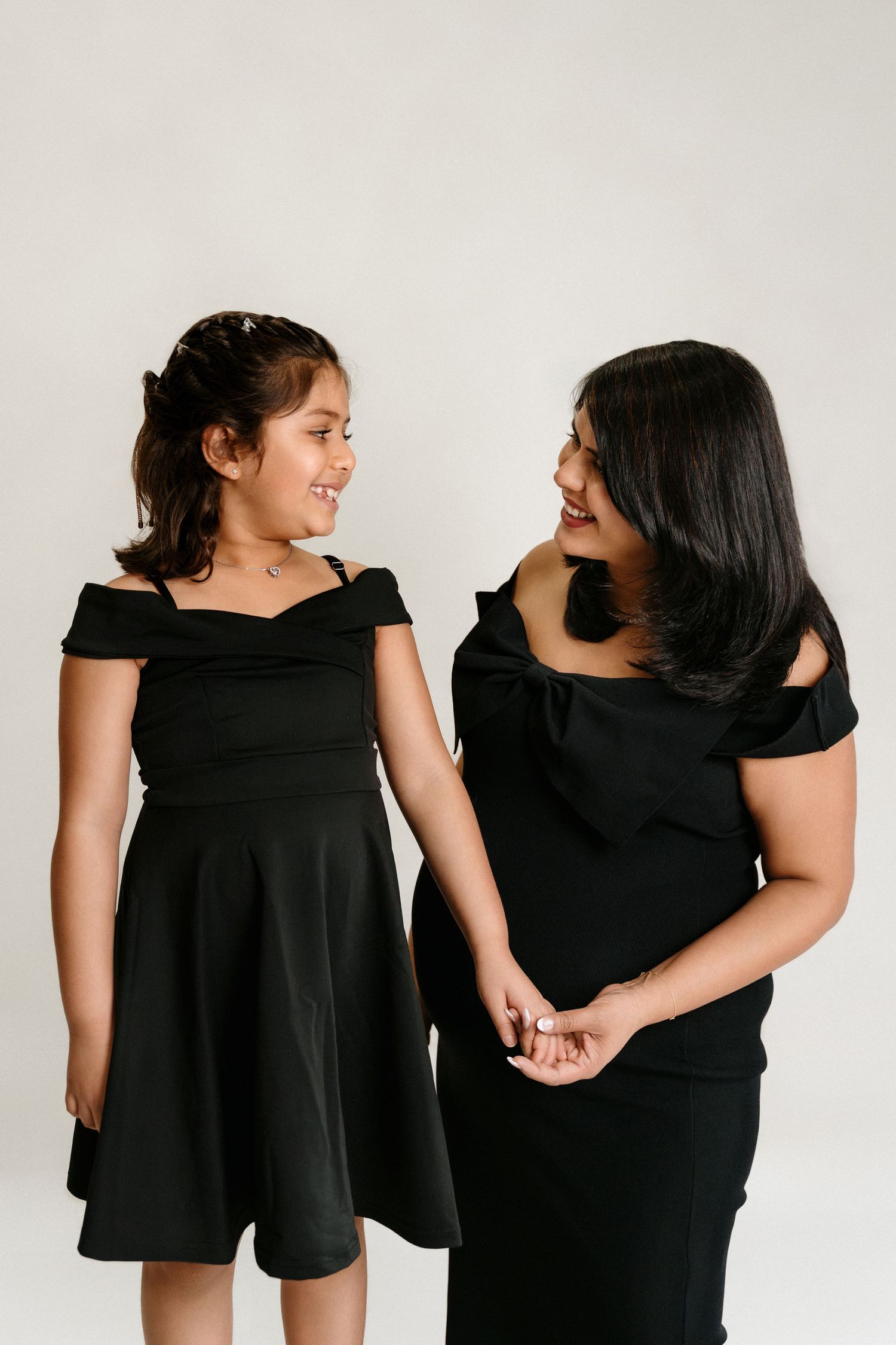 A woman and young girl in matching black off-shoulder dresses smile at each other while holding hands.