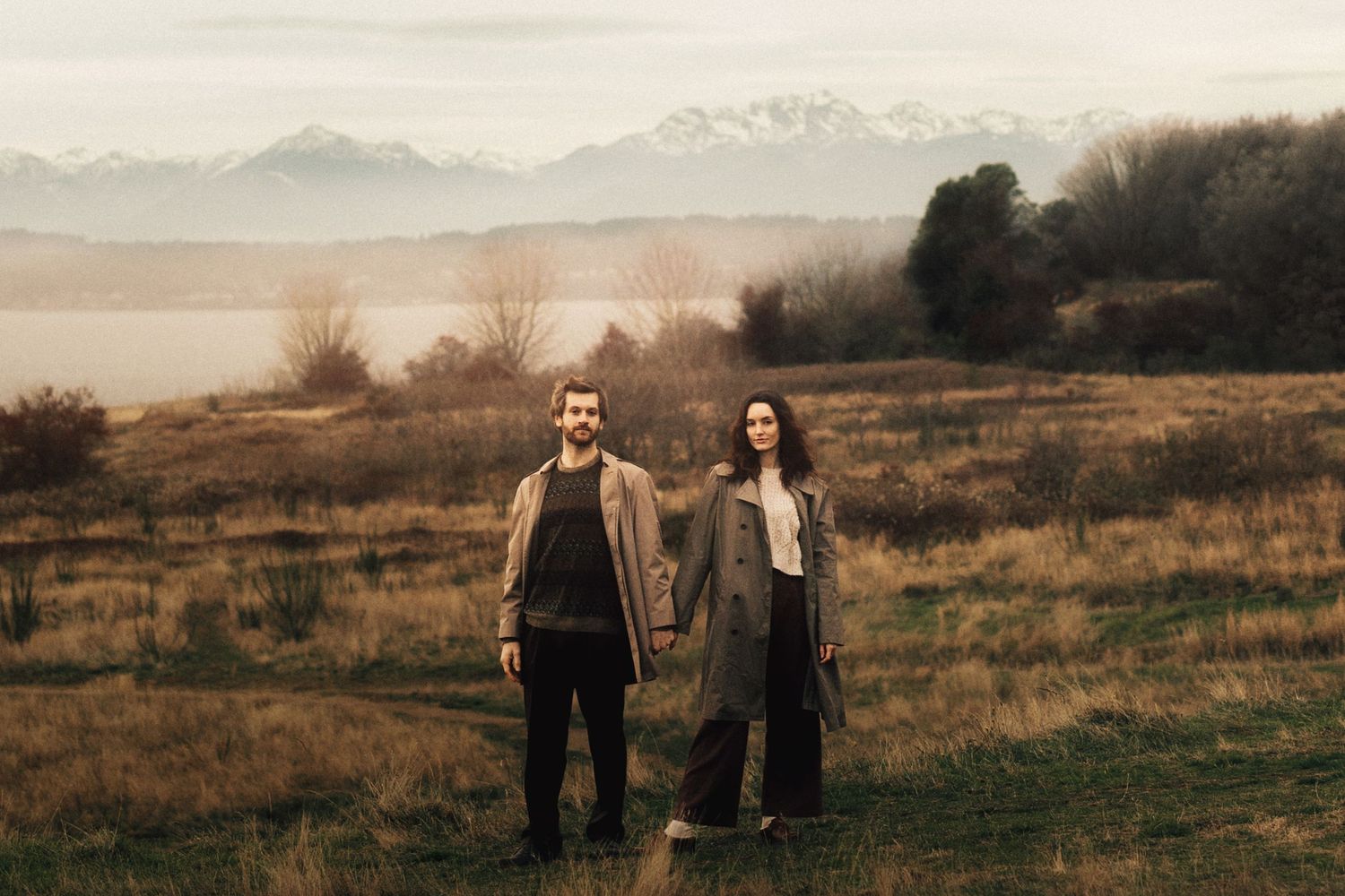 A couple stands in a misty field with snow-capped mountains and a lake in the background.