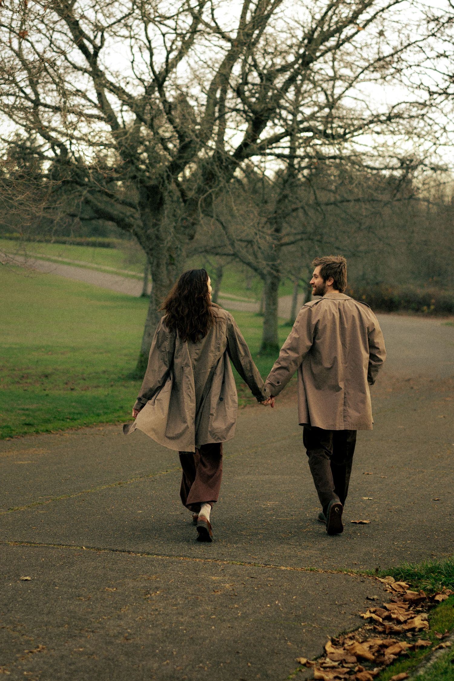 Couple strolling hand in hand on a park path surrounded by bare autumn trees, wearing matching trench coats.