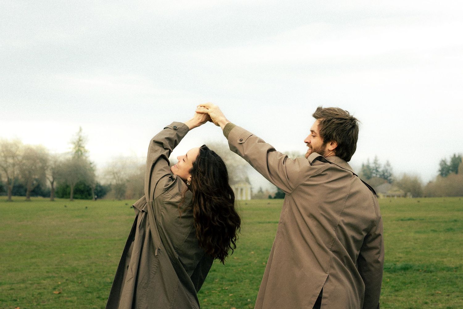 Couple dancing together in a green field on a cloudy day, both wearing matching brown coats.